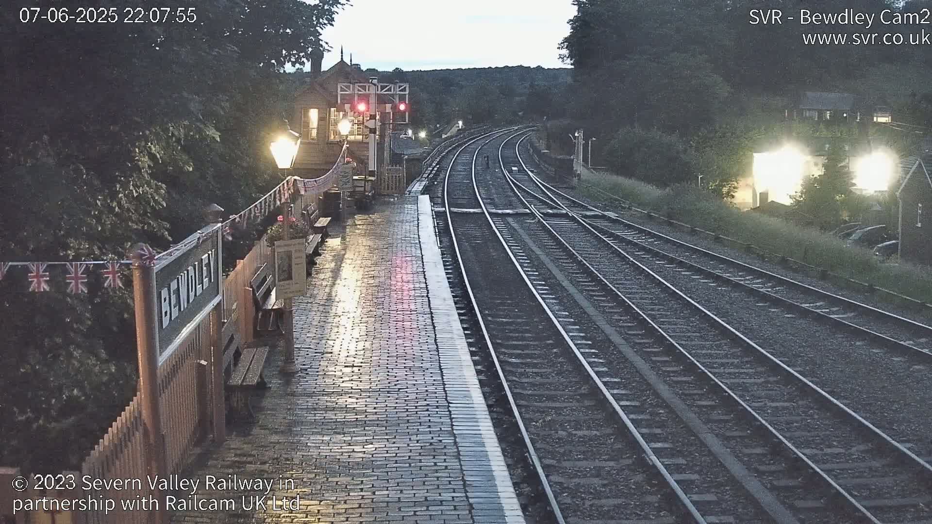A wet brick railway platform at twilight, with train tracks extending into the distance under a partly cloudy sky.