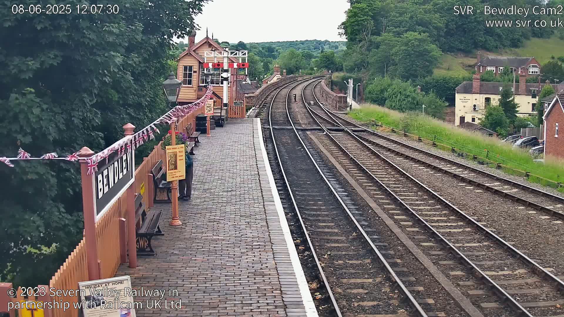A train station platform with two sets of tracks curving into the distance on a partly sunny day, surrounded by lush green hills and vegetation.