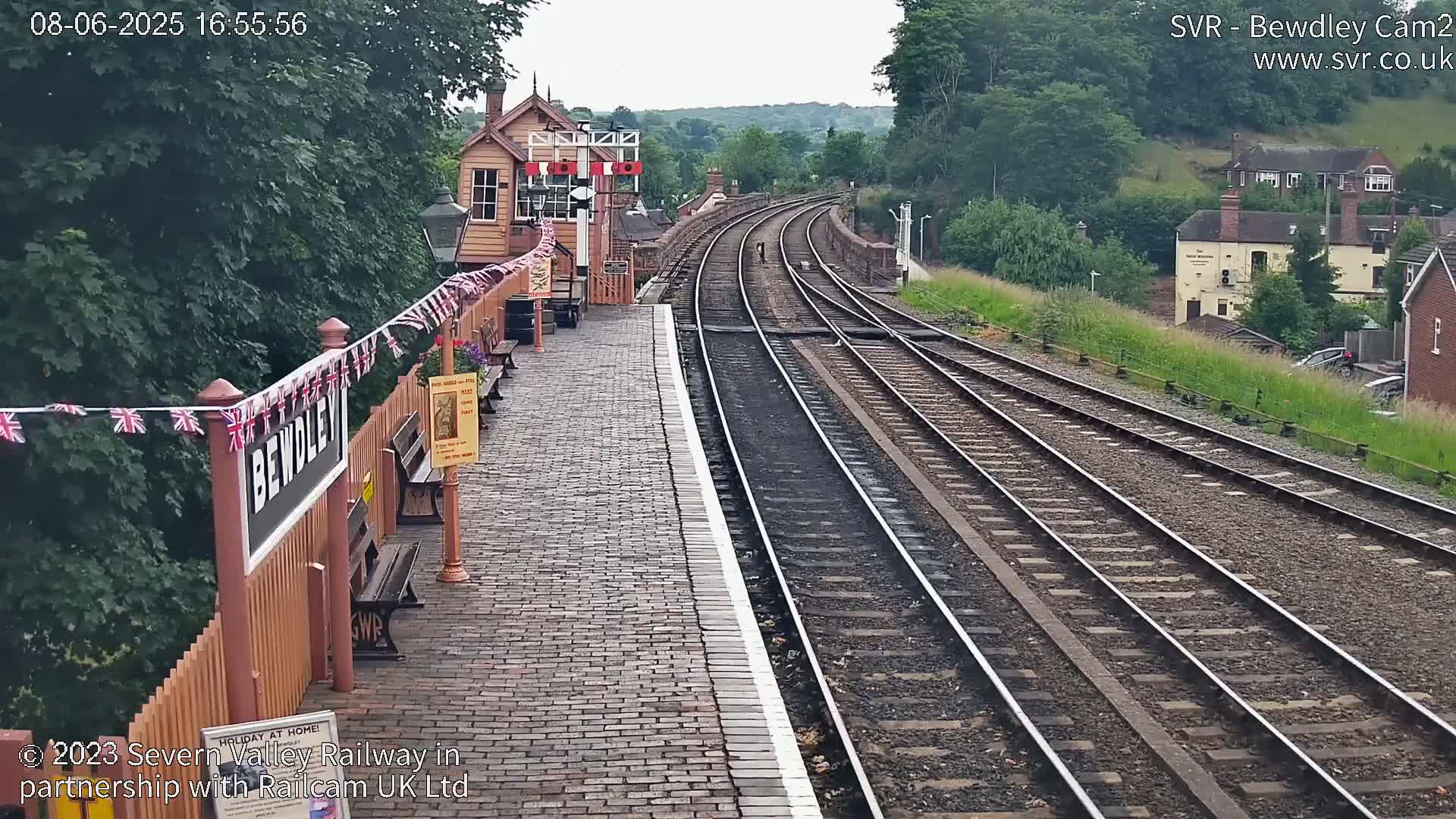 A train station platform with a brick surface and multiple train tracks curving into the distance on an overcast day, surrounded by lush green vegetation and buildings.