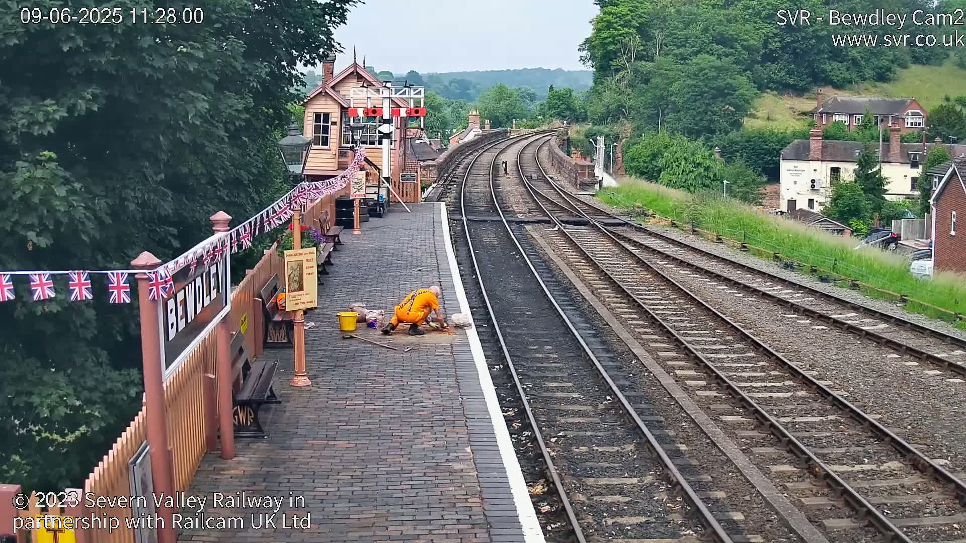 A person in orange work clothes performs maintenance on a brick railway platform beside two sets of train tracks that curve into the distance under a partly cloudy sky.
