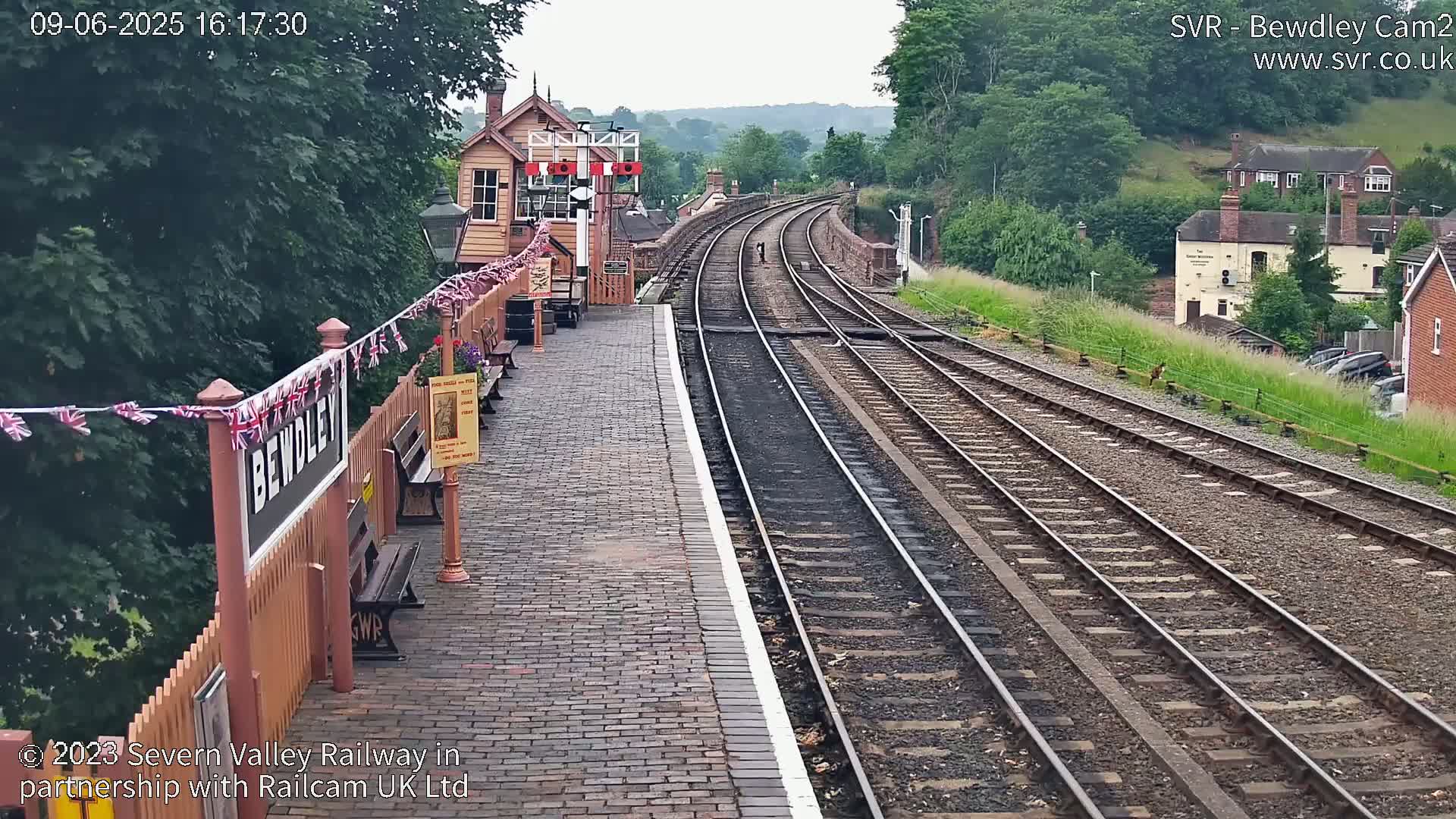 A train station platform with brick paving and a wooden fence, decorated with small flags, overlooks several railway tracks curving into the distance under an overcast sky.