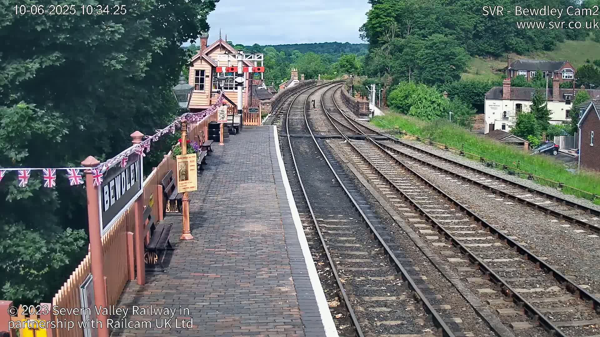 A train station platform with a brick surface, adorned with British flags, overlooks a curving railway track that extends into a hilly, green landscape under partly cloudy skies.