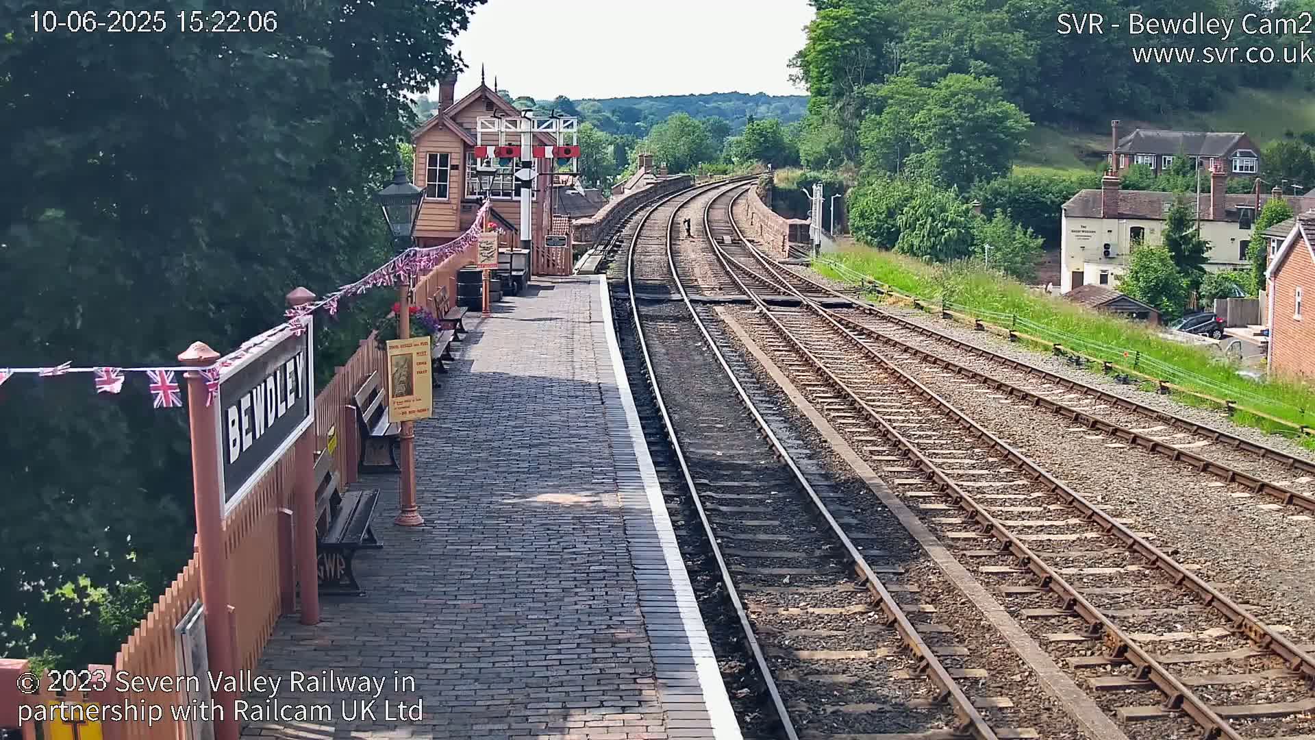 A sunny view of a train station platform with two sets of train tracks curving into the distance, bordered by a wooden fence and signal box.