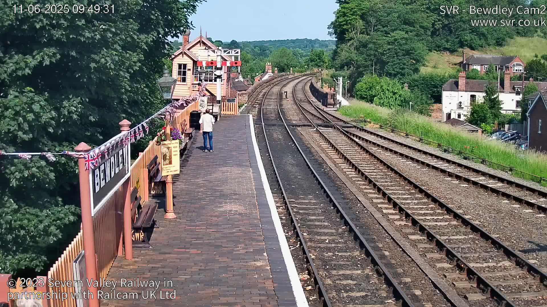 A sunny view of a train station platform with multiple train tracks curving into the distance, past buildings and lush green hills.