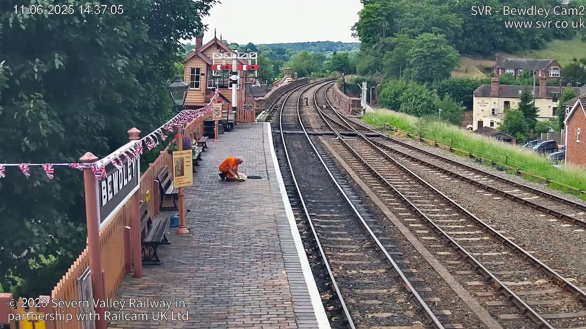 A person in an orange shirt kneels on a brick railway platform next to train tracks curving into the distance on a sunny day, with bunting strung along a nearby fence.