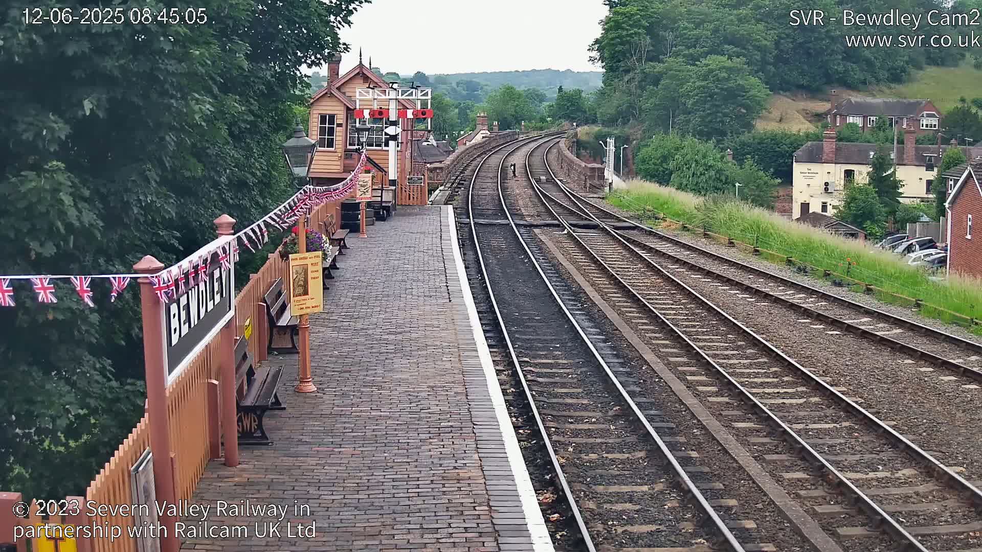 A train station platform with multiple train tracks extending into the distance on an overcast day, featuring a small signal box and bunting.