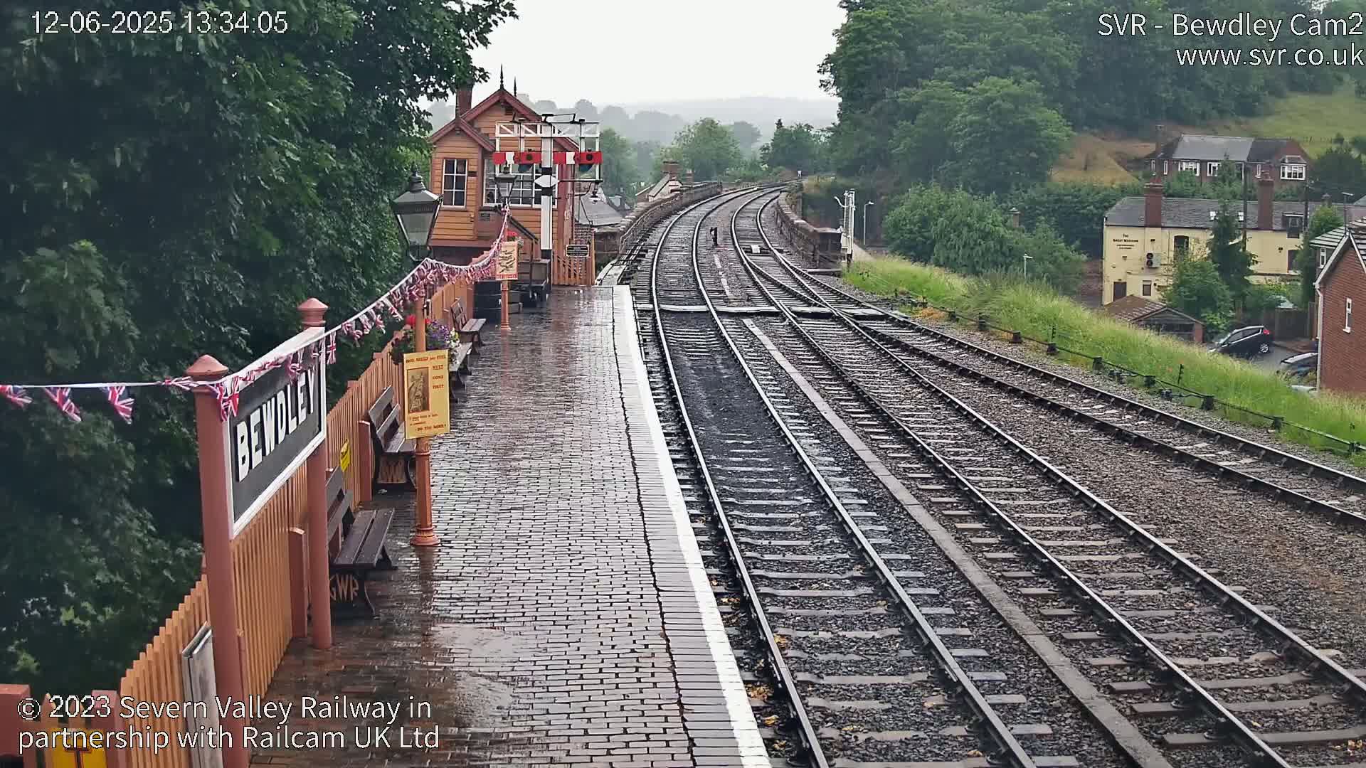 A wet train station platform with two sets of railway tracks extending into the distance under a rainy sky.