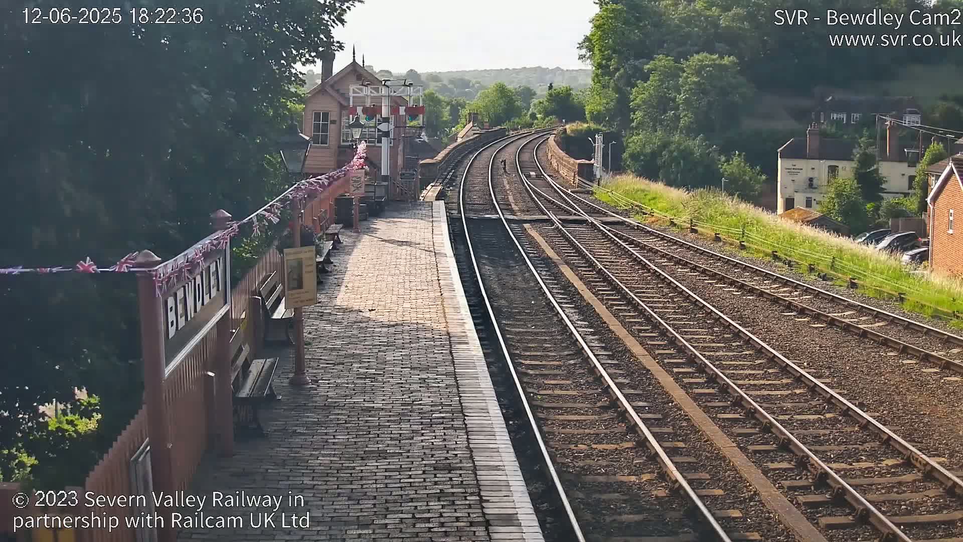 A sunny view of a train station platform and railway tracks extending into a wooded hillside.