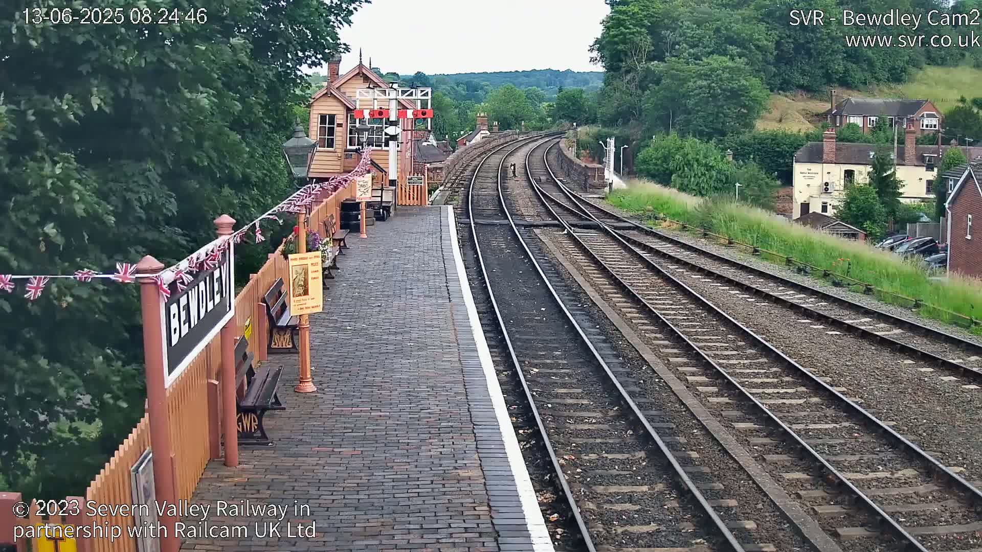 A brick railway platform with a signal box and multiple train tracks curving into the distance on an overcast day, bordered by lush greenery and buildings.