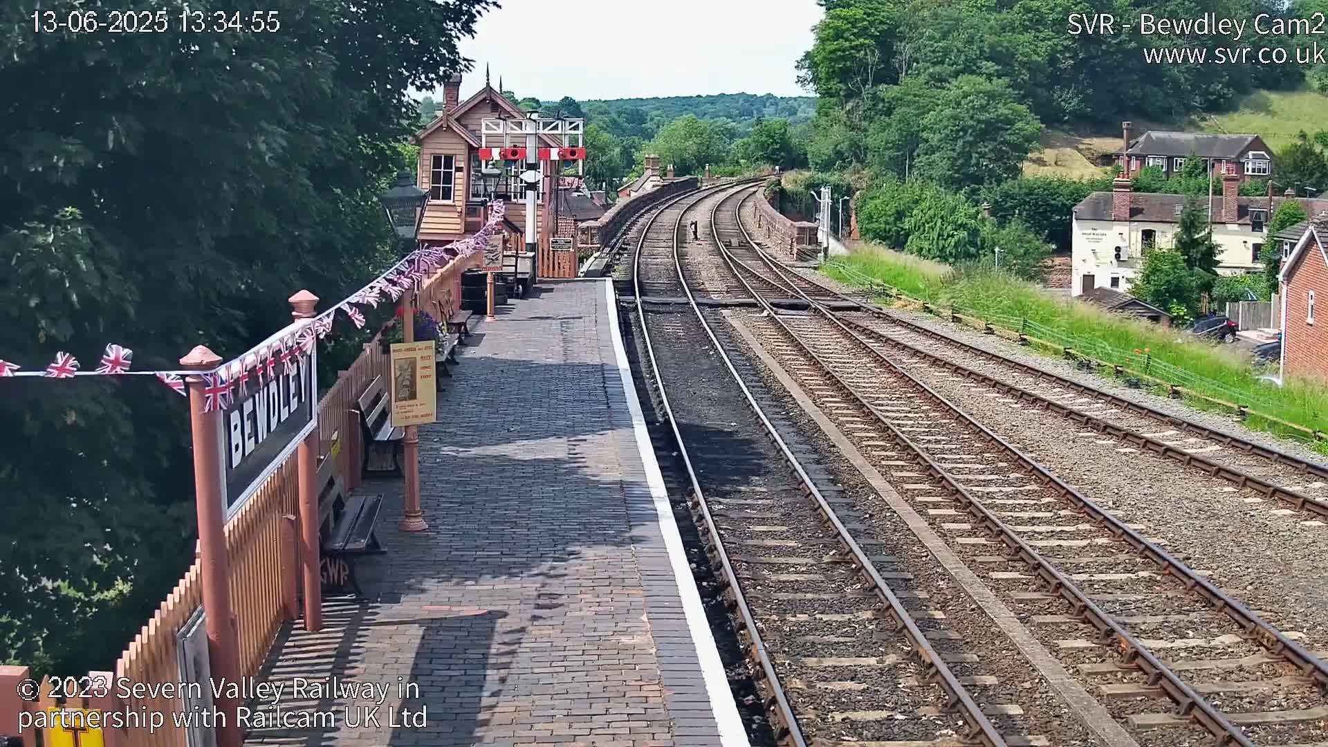 A sunny view of a train station platform with multiple train tracks extending into the distance, alongside a wooden fence decorated with bunting.
