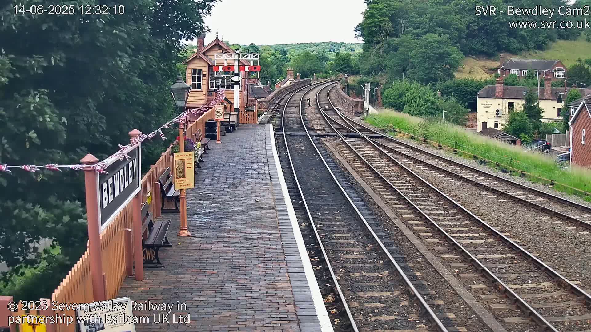 A train station platform with a brick pavement and multiple train tracks extending into a verdant hillside under a partly cloudy sky.