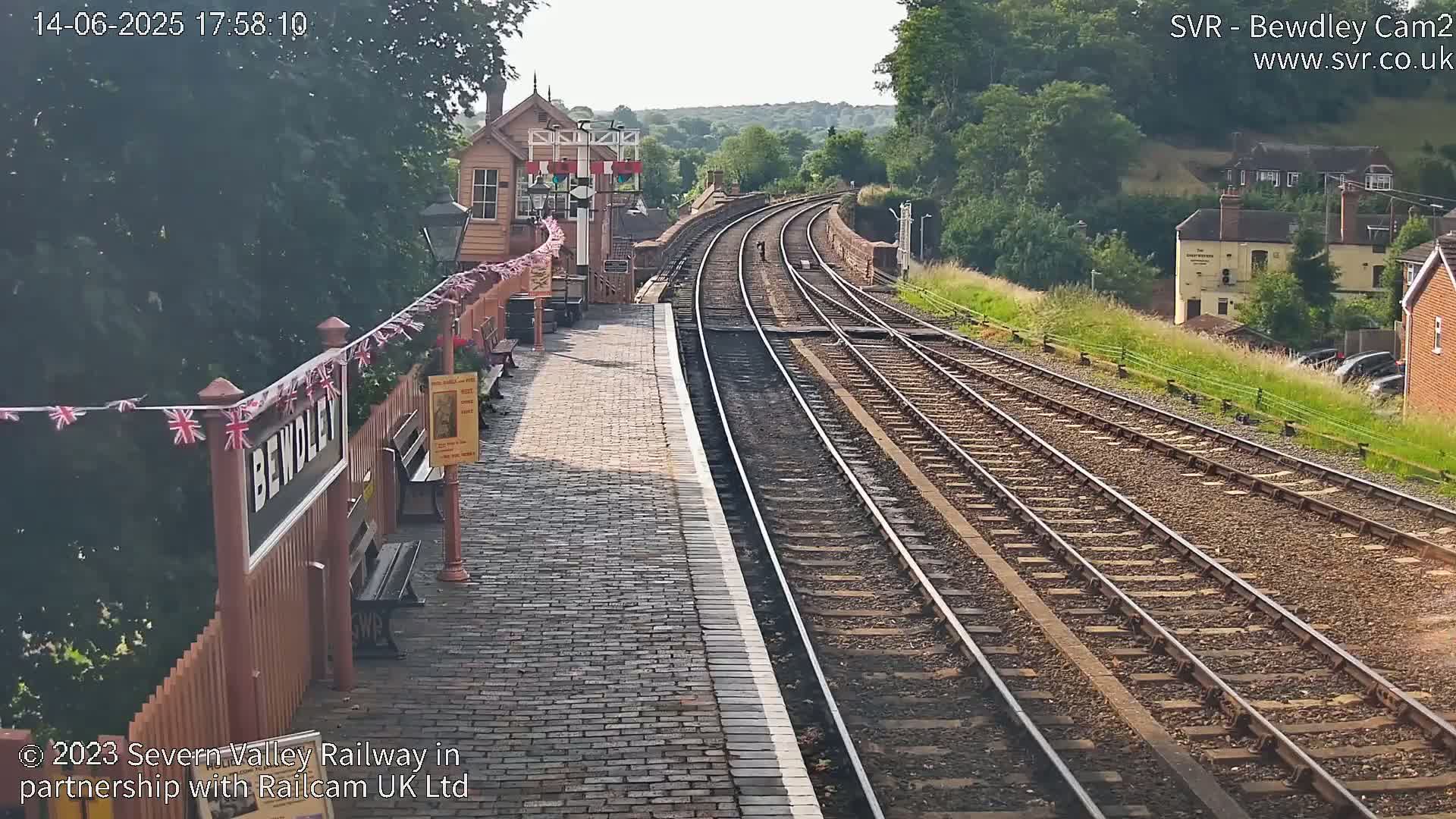 A train station platform with brick paving and multiple train tracks curving into the distance on a sunny day, surrounded by greenery and buildings.