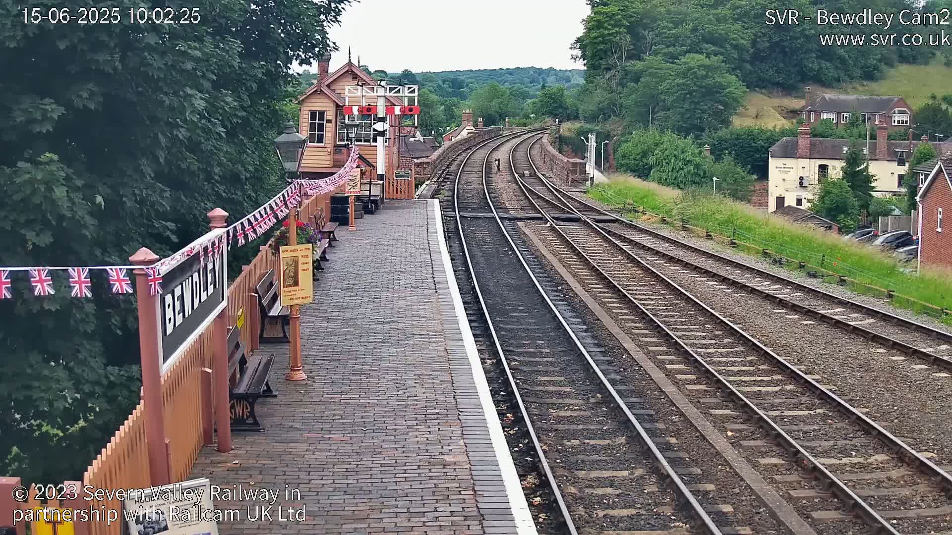 A train station platform with a brick pavement and multiple train tracks curving into the distance on a partly sunny day, with lush green foliage surrounding the area.