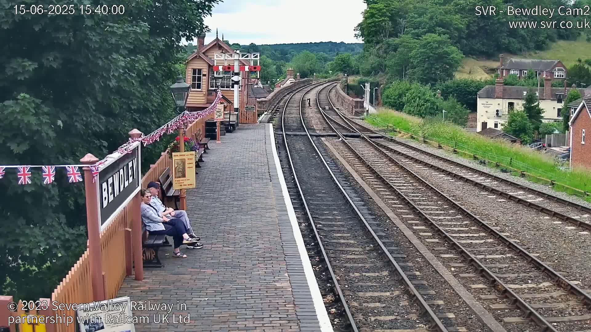 A brick train platform with two seated people overlooks a pair of railway tracks curving into a green, tree-lined hillside under a partly sunny sky.