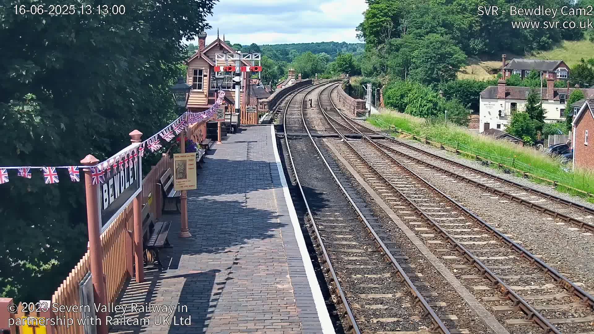 A sunny day at a train station with a brick platform and multiple train tracks extending into the distance, surrounded by greenery and buildings.