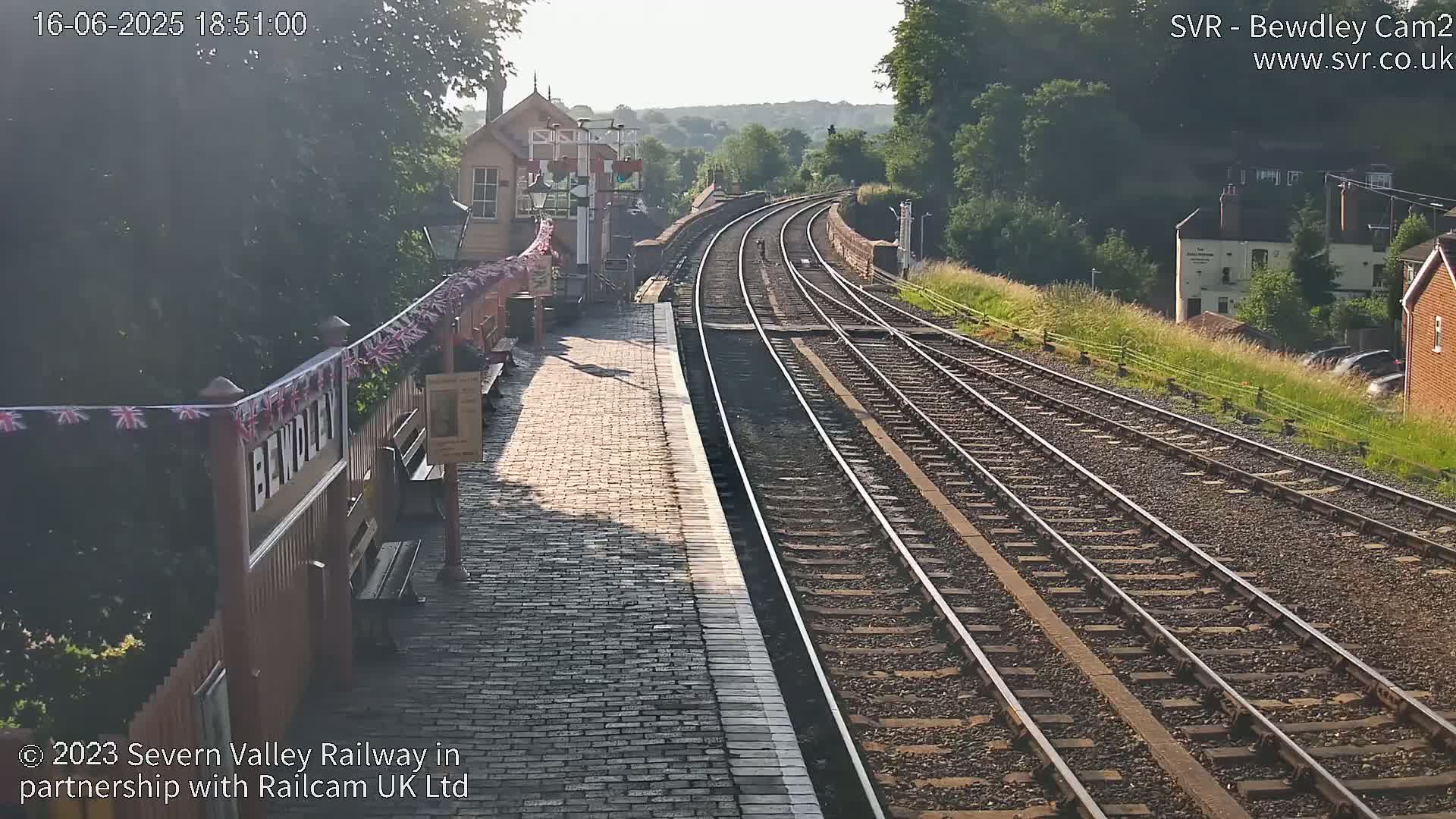 A sunny view of a railway station platform and tracks, extending into a tree-lined hillside in the distance.