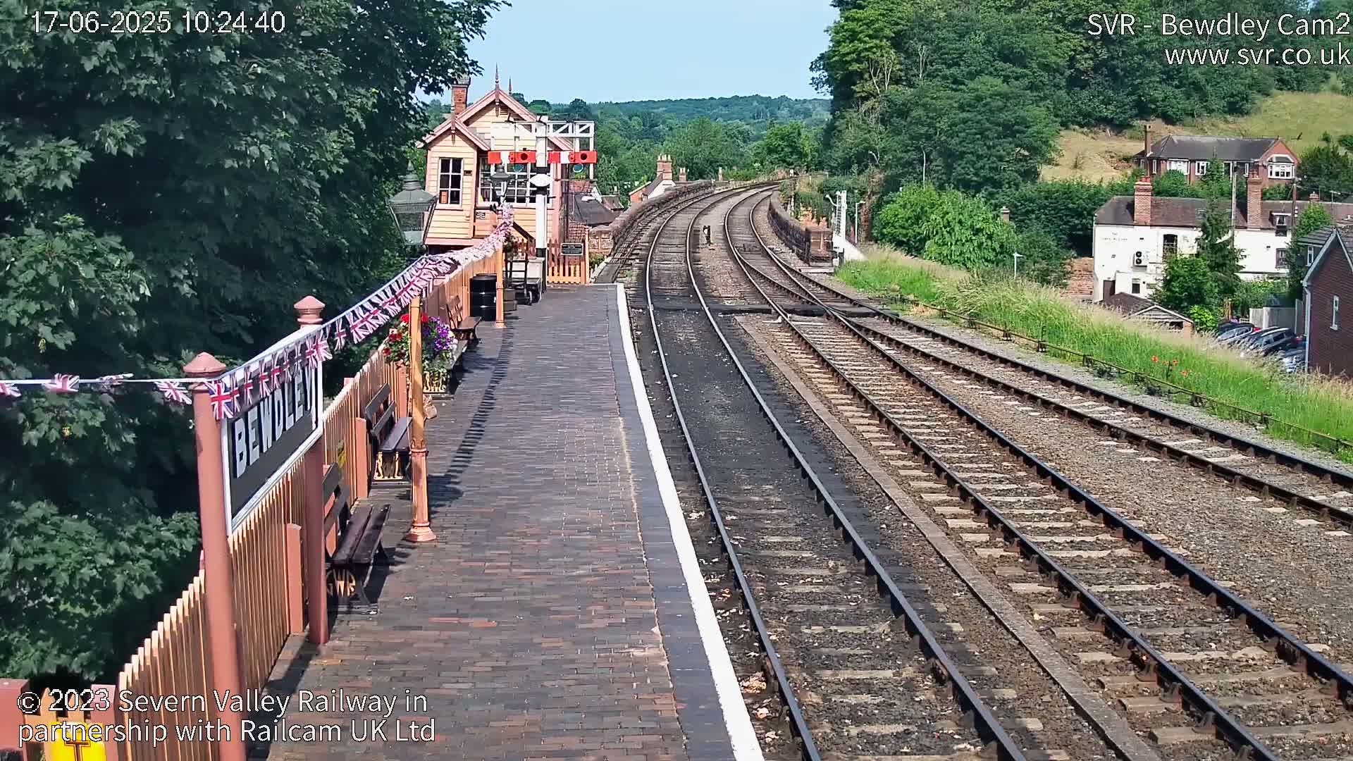 A sunny view of a train station platform and railway tracks leading into a wooded hillside.