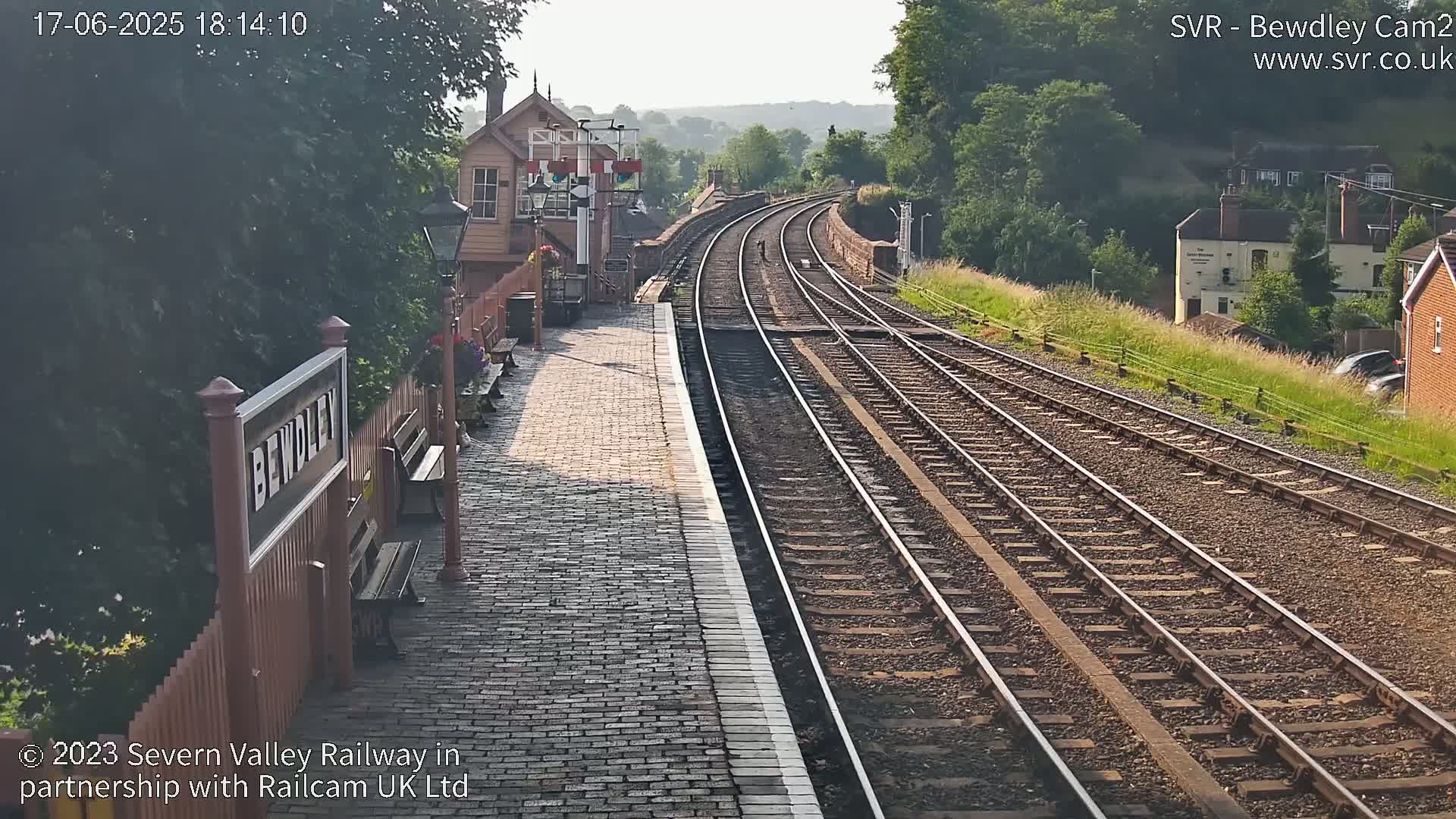 A train station platform with brick paving and several benches overlooks two sets of train tracks curving into the distance under a sunny sky.