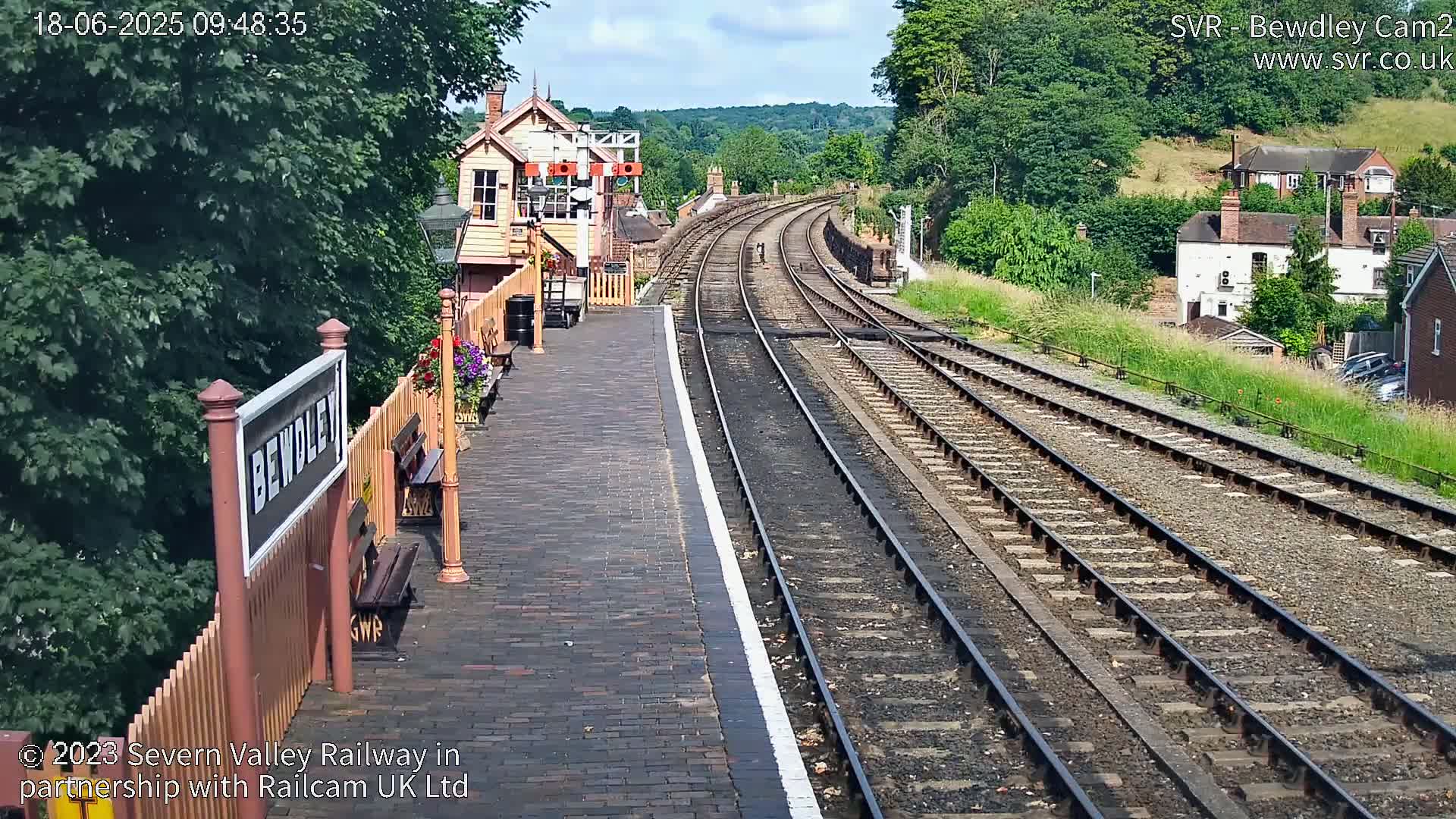 A train station platform on a sunny day, with railway tracks extending into the distance, surrounded by lush greenery and buildings.