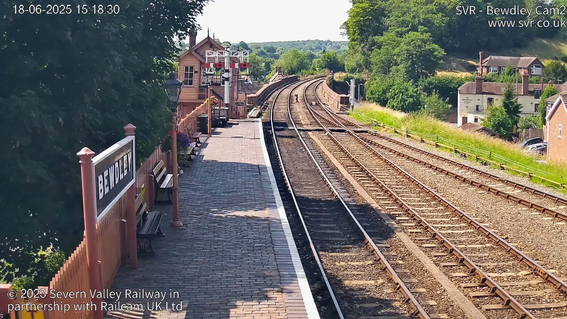 A sunny view of a train station platform with two sets of railway tracks curving into the distance, lined with greenery and buildings.