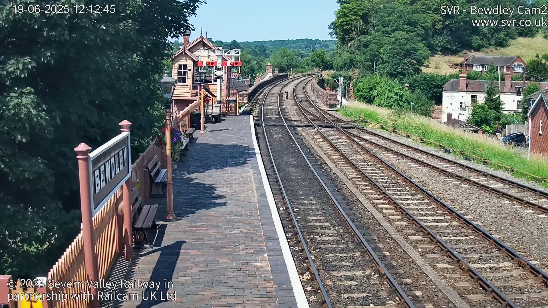 A sunny day at a train station with a brick platform and two sets of railway tracks curving into the distance, surrounded by green trees and buildings.