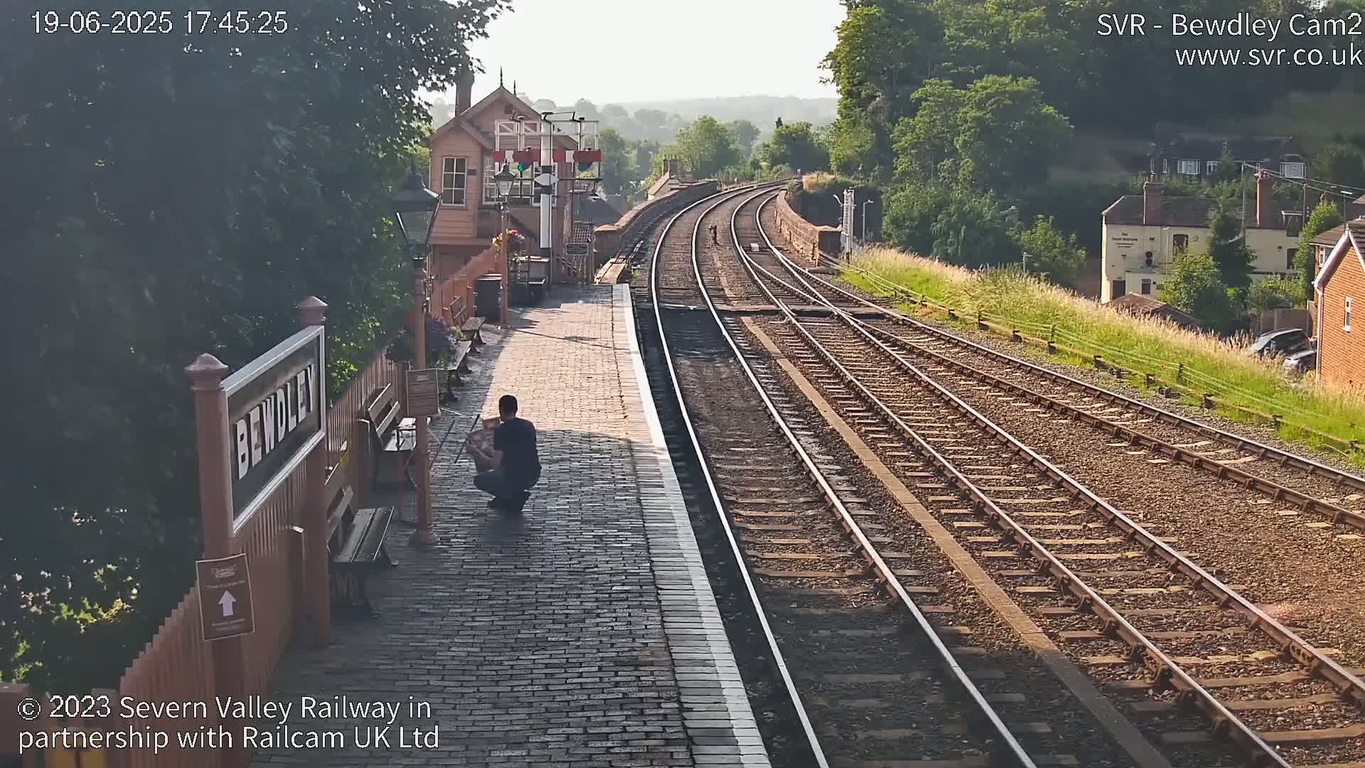 A person sits on a brick platform beside train tracks that curve into the distance under a sunny sky.