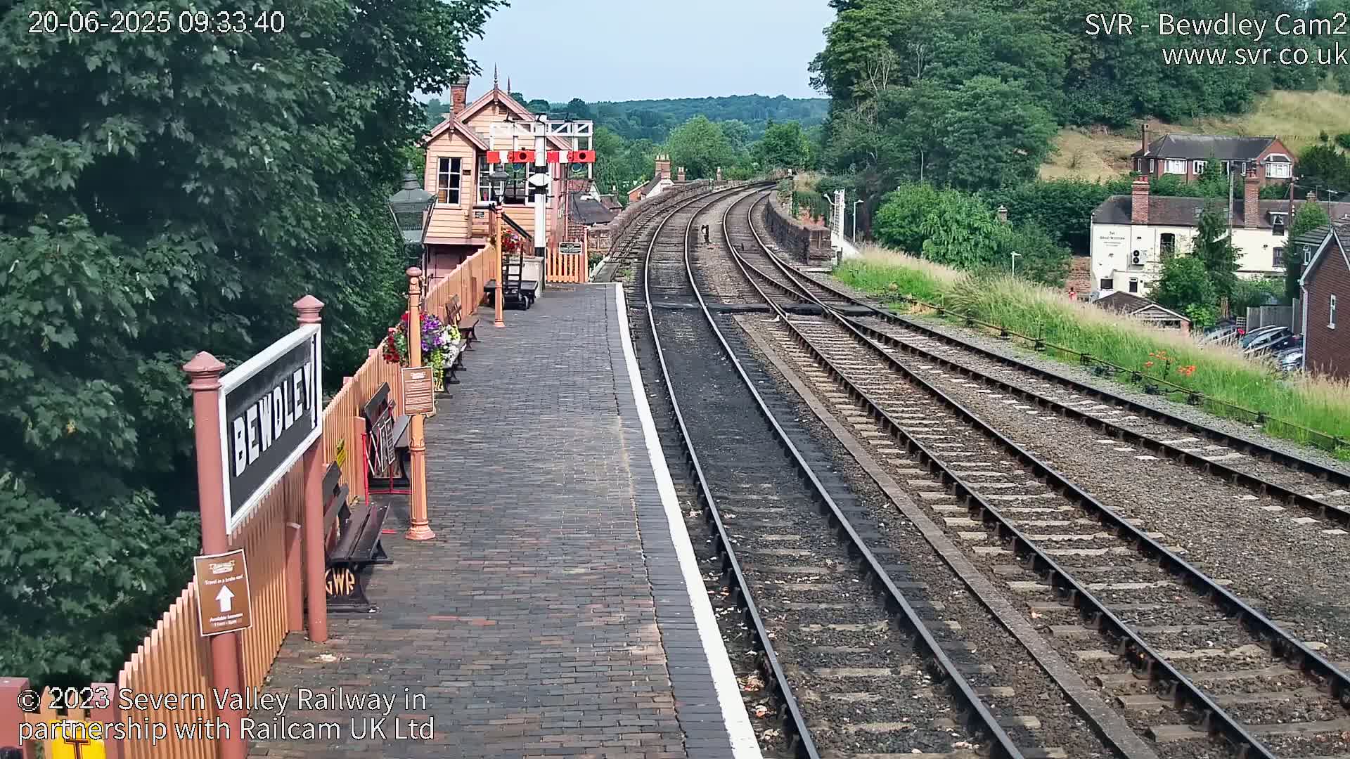 A brick railway platform with a signal box and multiple train tracks extending into a distance under a partly sunny sky.