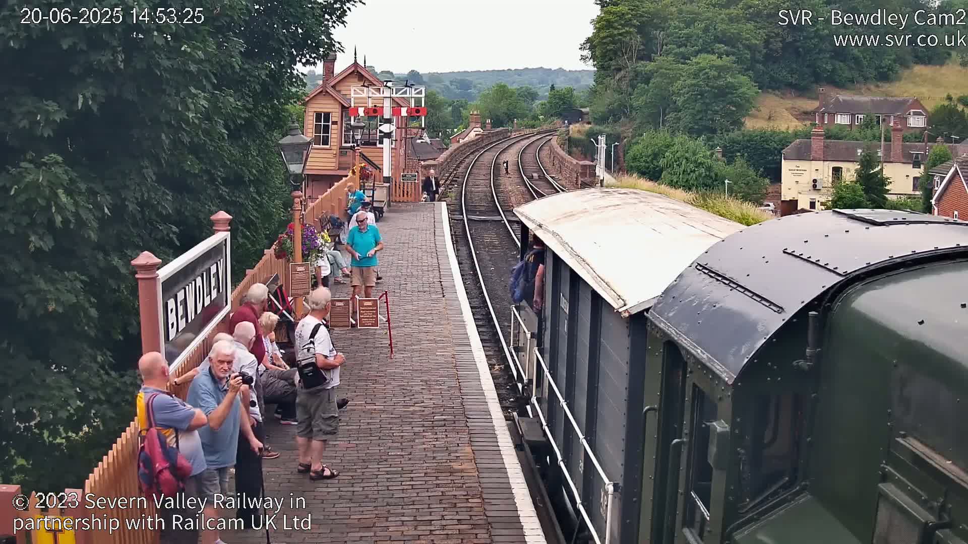A train pulls into a brick railway station on a partly sunny day, with several people waiting on the platform.