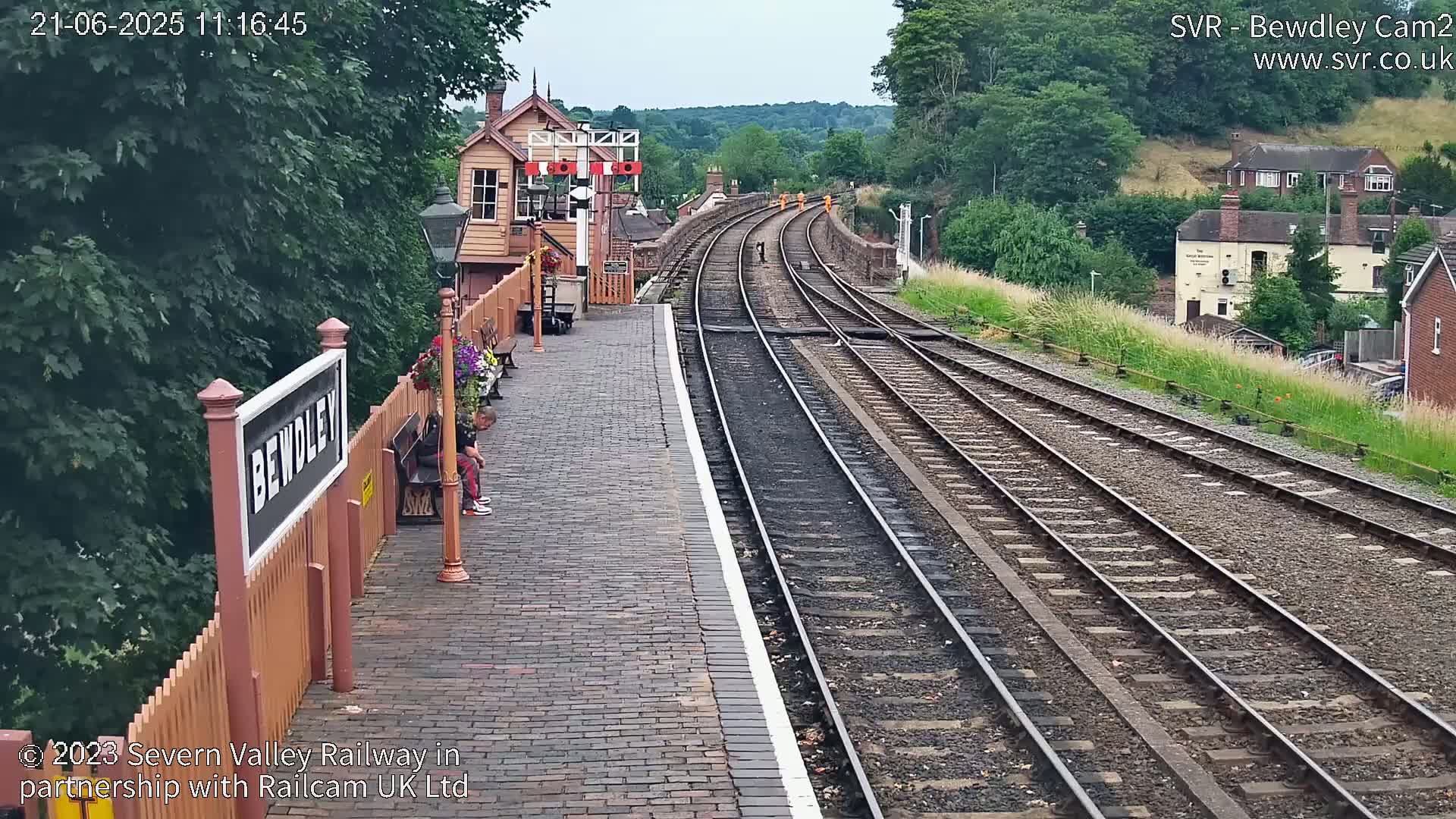 A brick train platform with a wooden fence and signal box overlooks a railway track curving into the distance under a partly cloudy sky.
