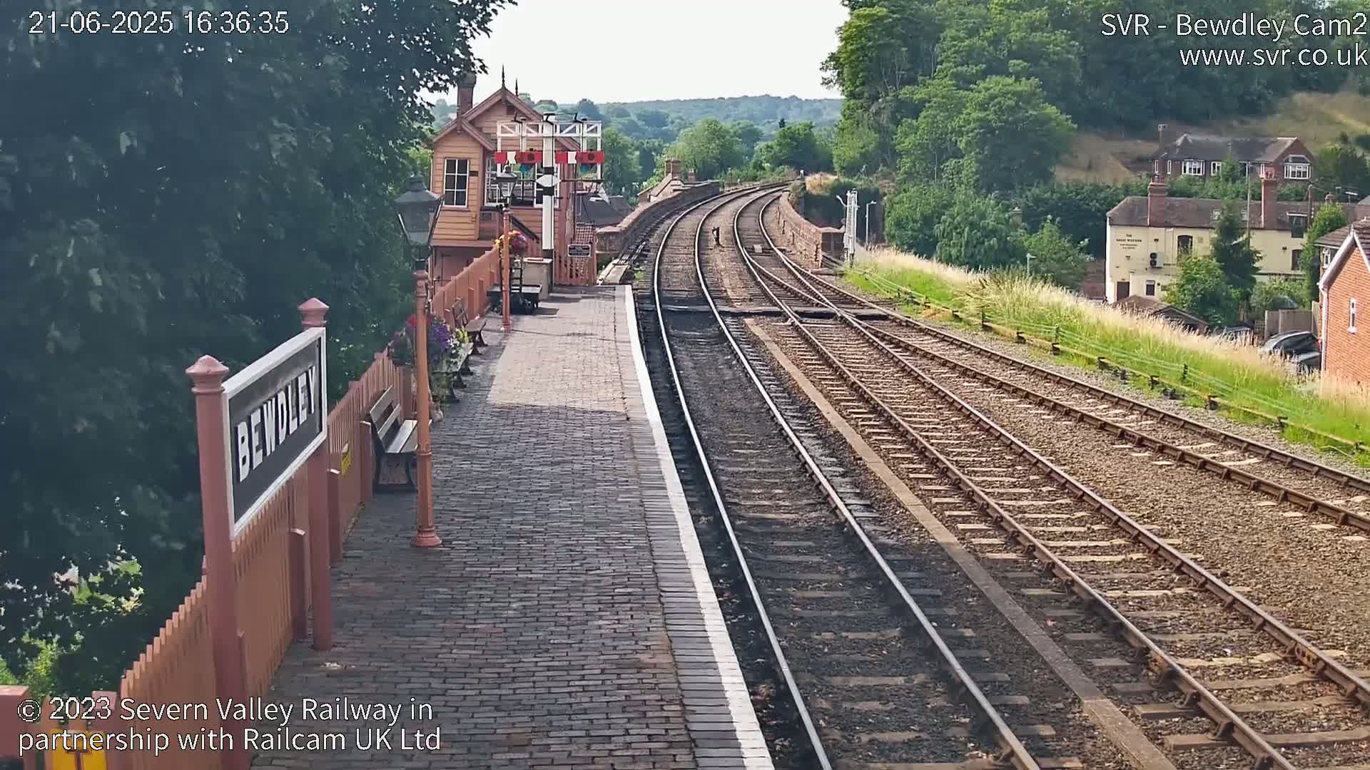 A brick railway platform with a signal box and curving train tracks extends into a sunny, tree-lined hillside.