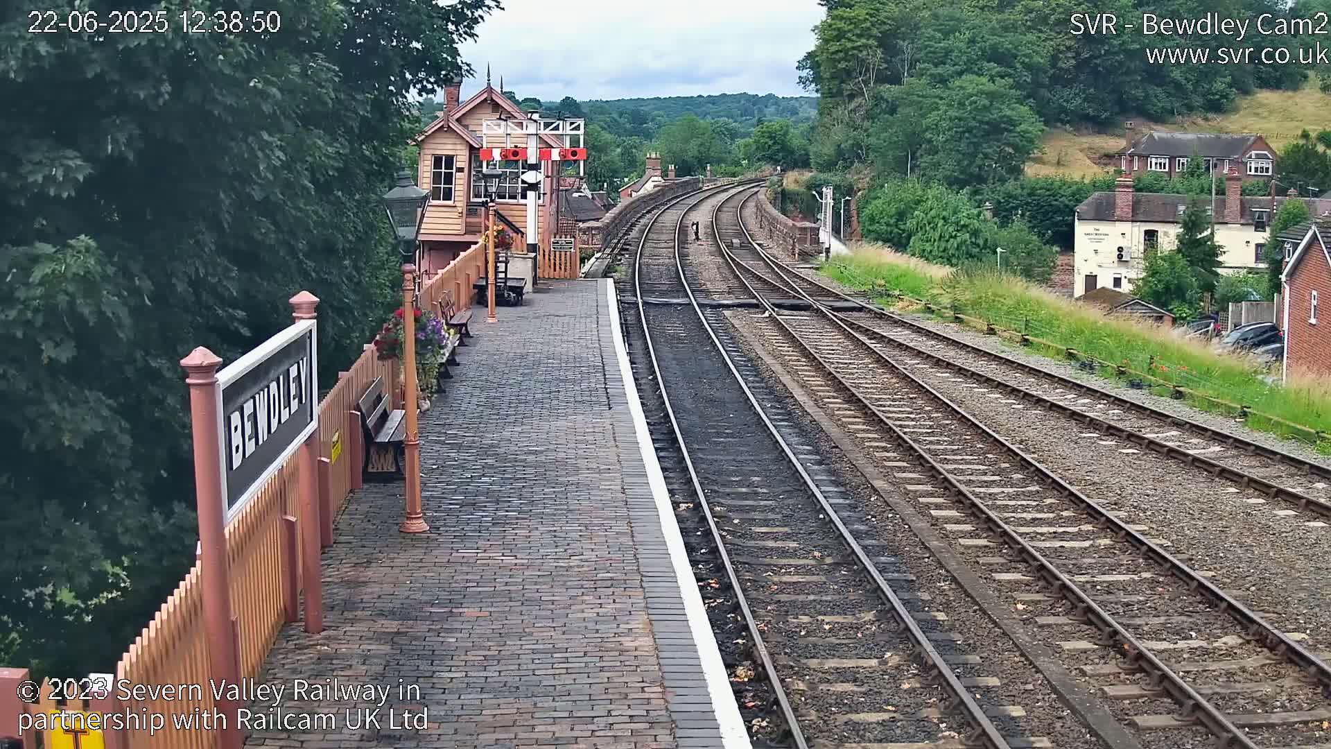 A train station platform with a brick pathway next to several train tracks curving into the distance under a partly cloudy sky.