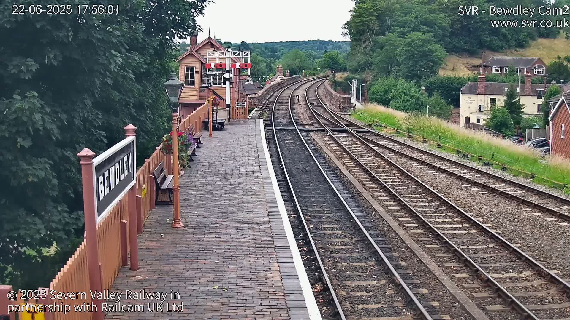 A train station platform with brick paving and a signal box, overlooking curving railway tracks extending into a verdant hillside under a partly cloudy sky.
