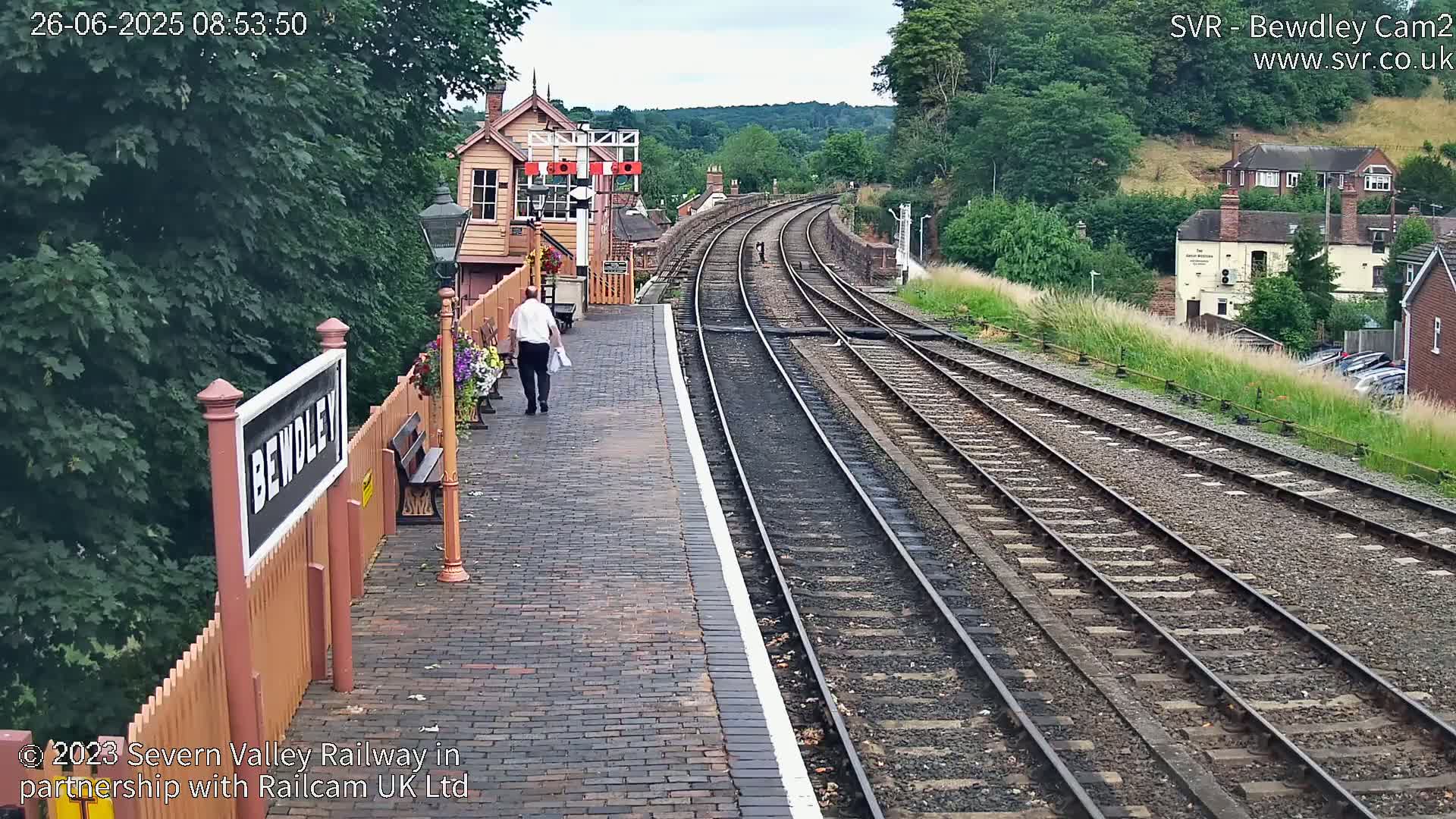 A person walks away from the camera along a brick train platform towards a small wooden building, with multiple train tracks curving into the distance under an overcast sky.