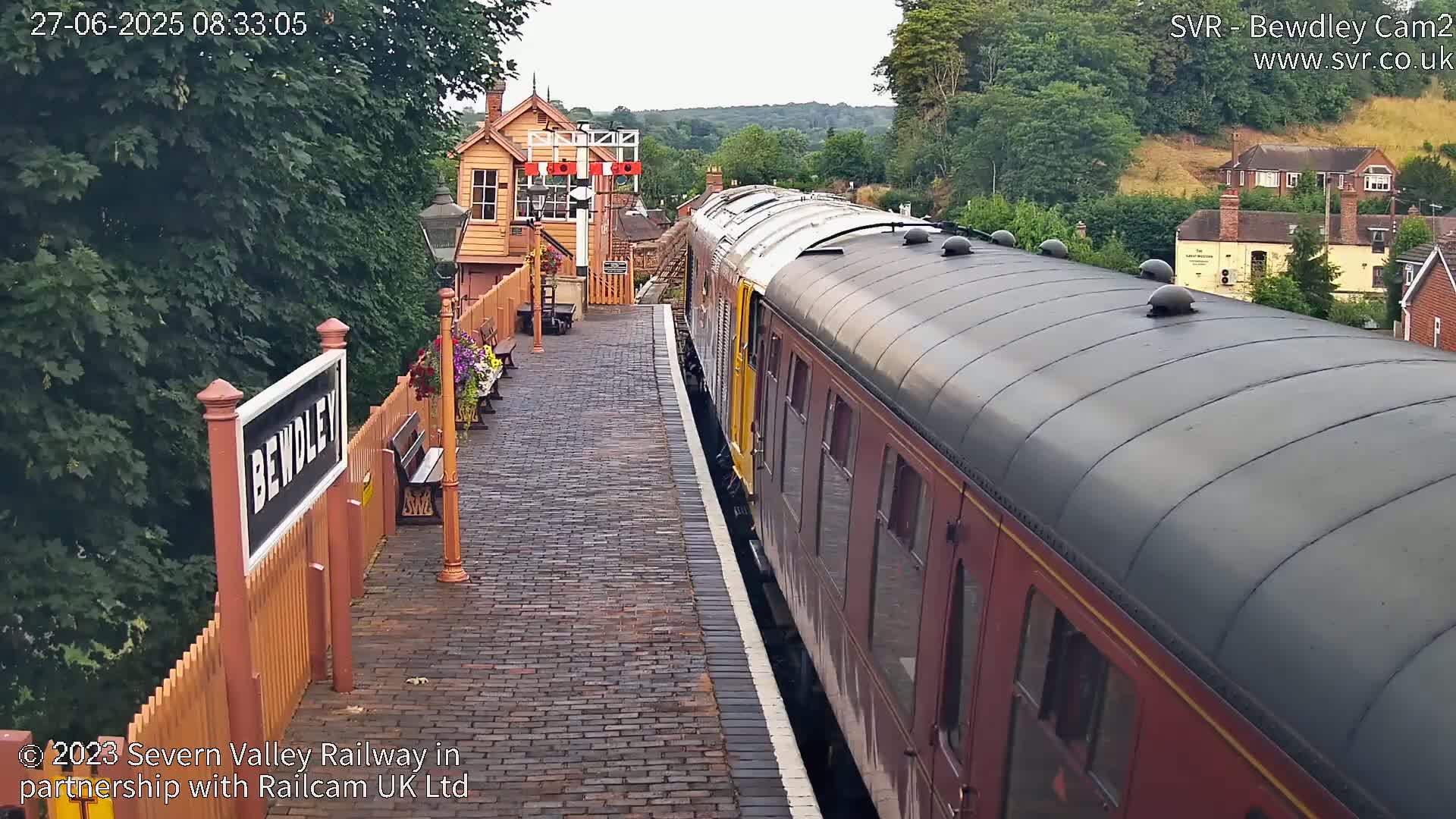 A maroon passenger train is stopped at a brick station platform on an overcast day.