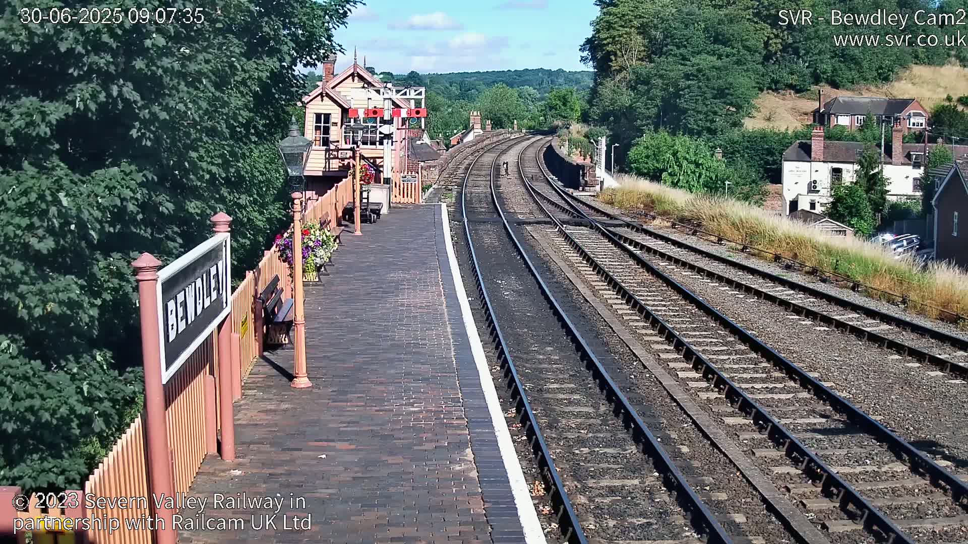 A sunny view of a train station platform with railway tracks extending into the distance, bordered by greenery and buildings.