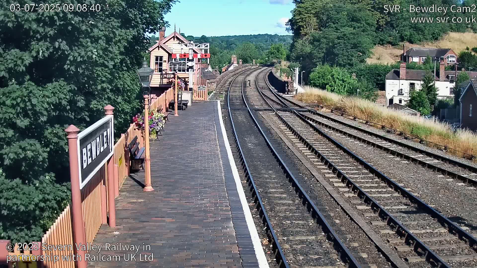 A sunny view of a train station platform with a railway extending into the distance, surrounded by green hills and trees.