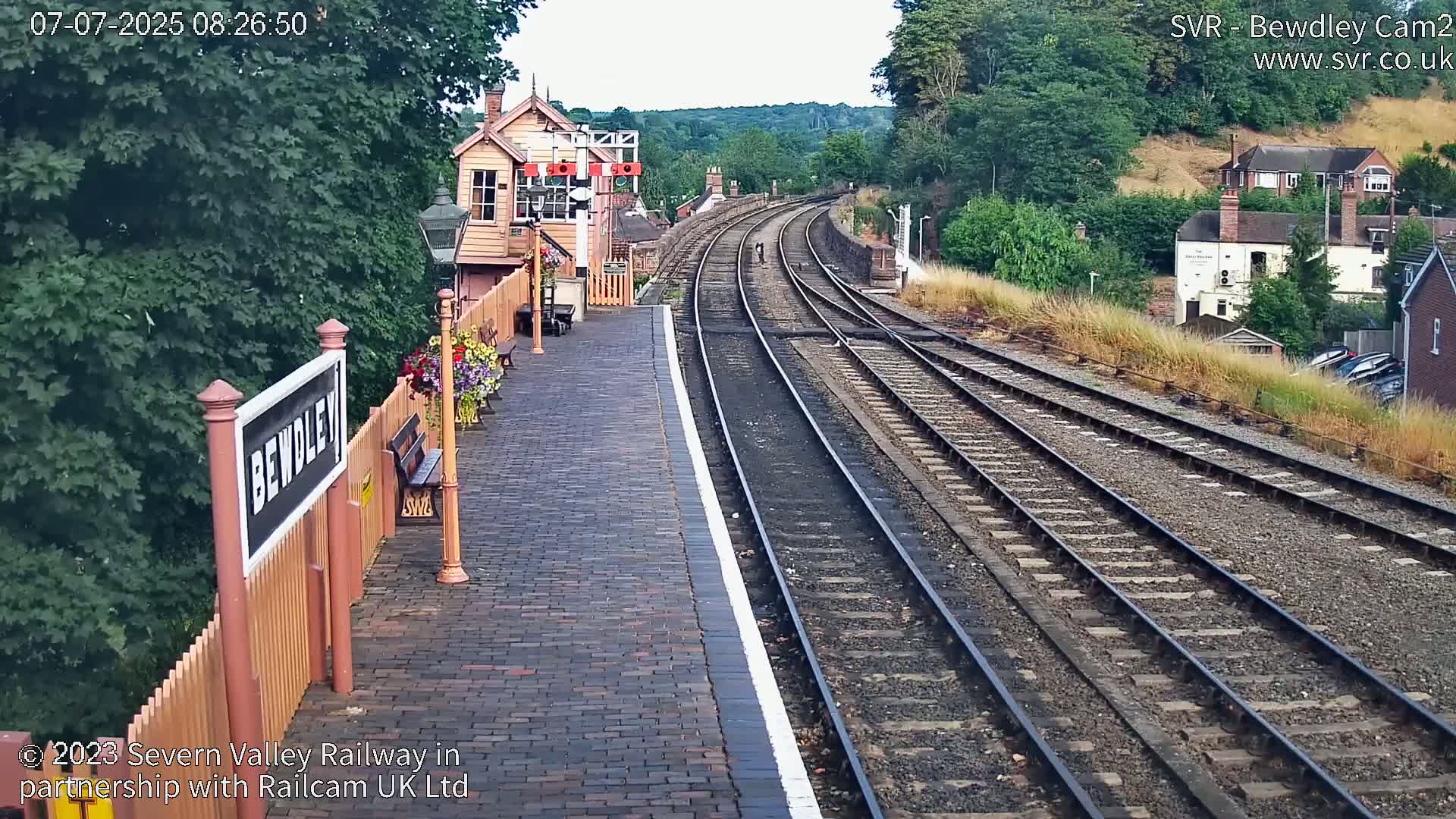A brick railway platform with a signal box and curving tracks extends into a hilly, tree-lined landscape on a partly sunny day.