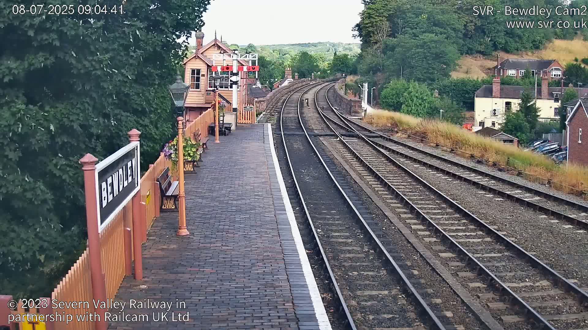 A brick train platform with a station building and multiple train tracks curving into the distance on a partly sunny day.