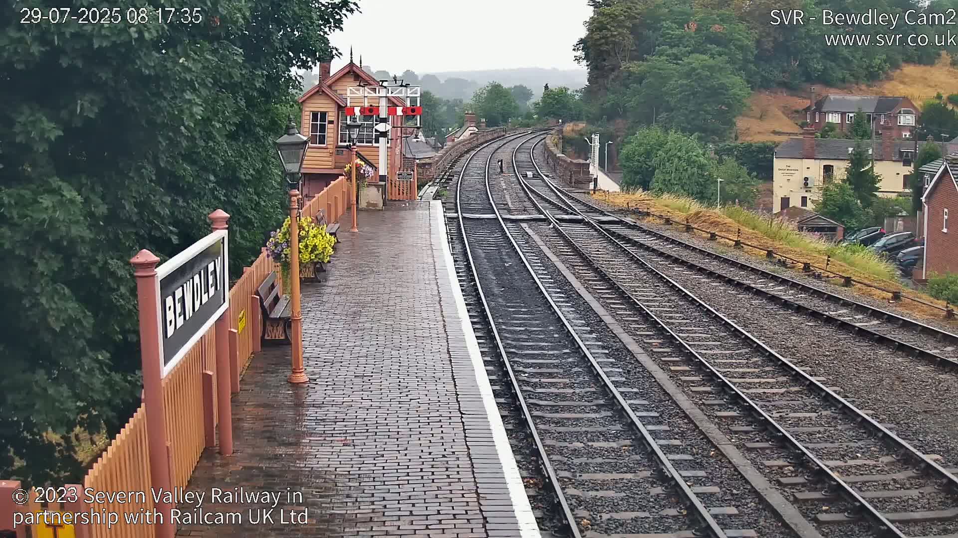 A wet, brick-paved train platform shows two sets of train tracks curving into the distance under an overcast sky.