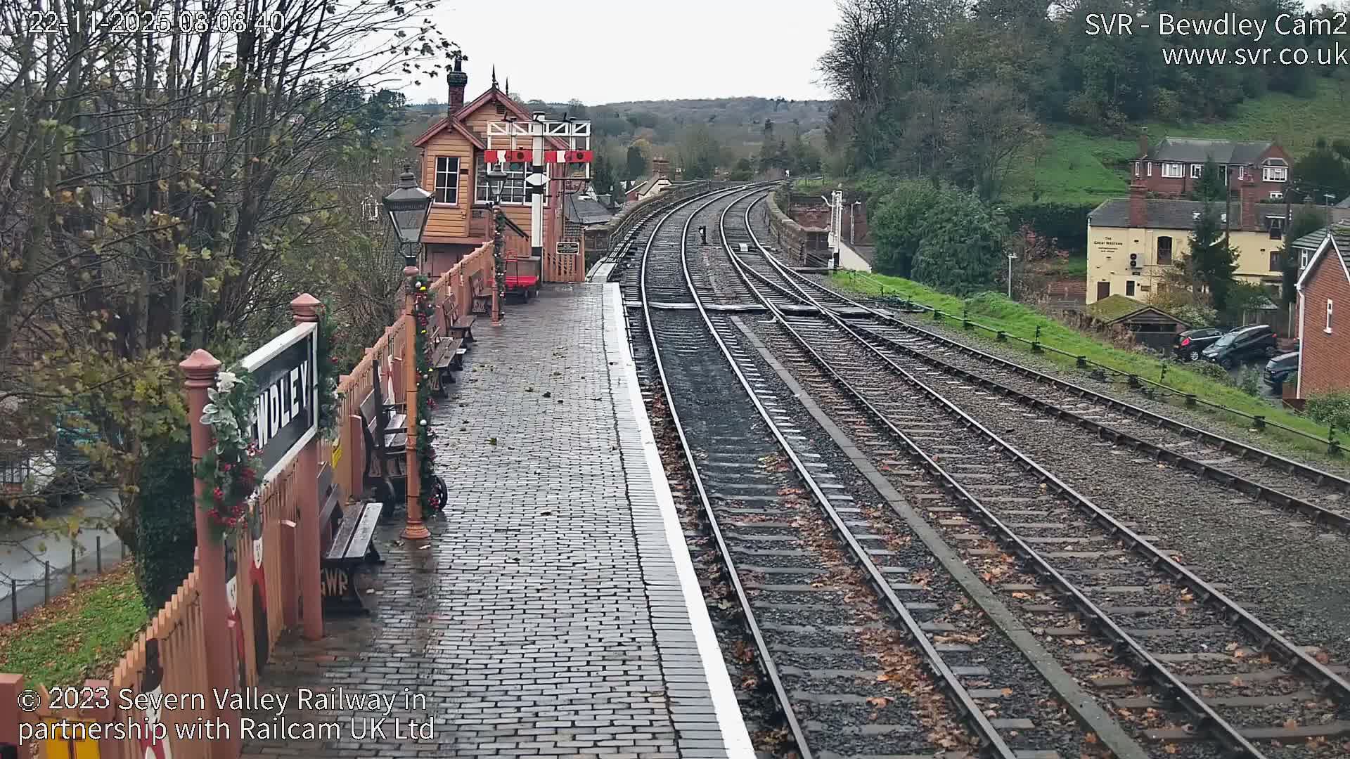 A festive, historic railway station platform, adorned with Christmas decorations and a wooden signal box, overlooks multiple curving tracks under a cloudy sky with scattered autumn leaves.