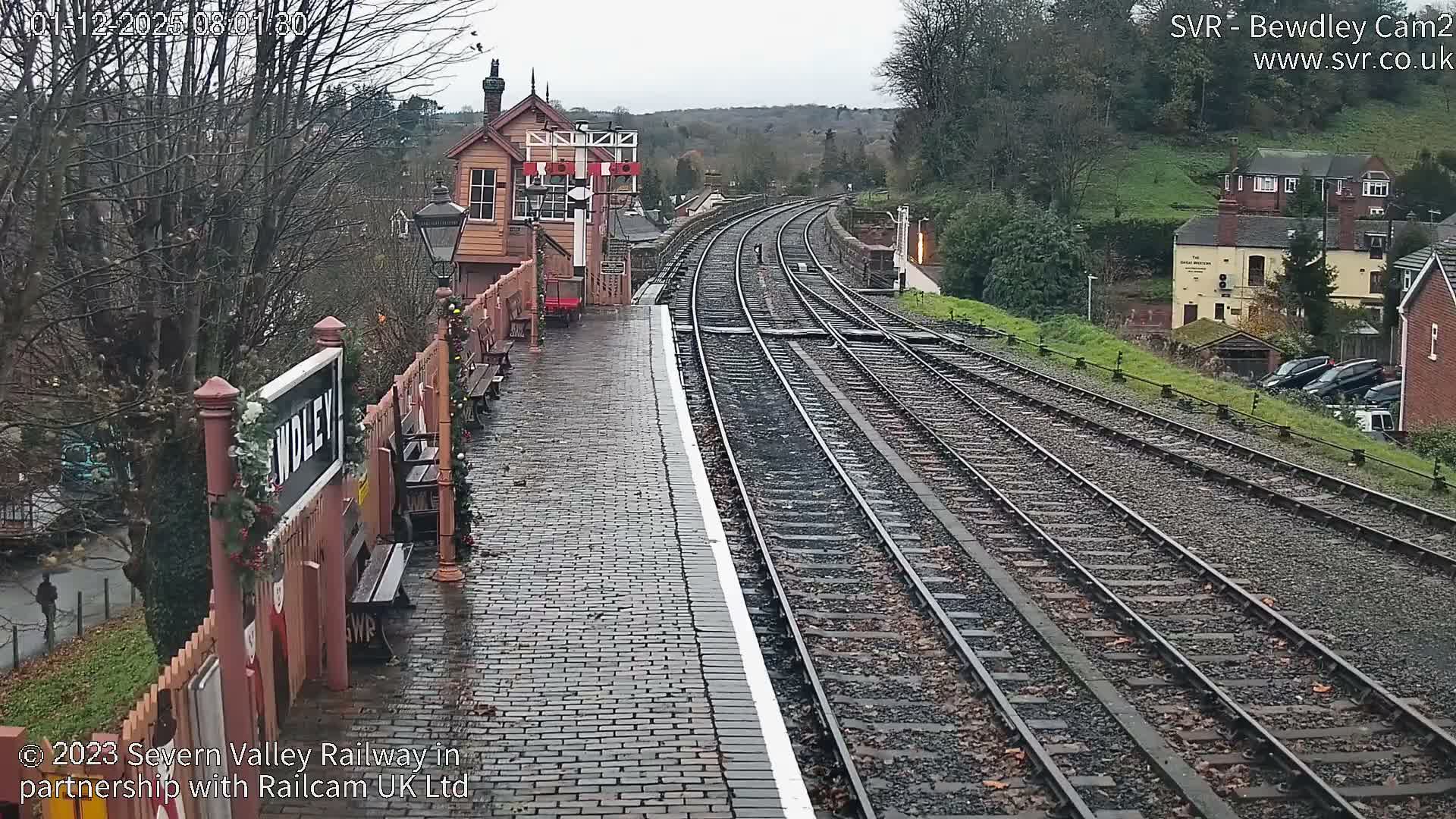 On an overcast and damp winter day, a historic train station features a cobblestone platform adorned with festive garlands, multiple curving tracks, and a classic signal box, all set against a backdrop of bare trees and distant hills.