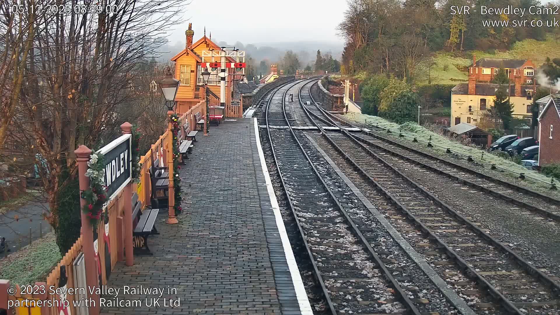 A charming old-fashioned railway station platform, adorned with festive garlands and benches, stretches alongside multiple frosty tracks that curve into a misty, tree-lined landscape under a cold, hazy sky.