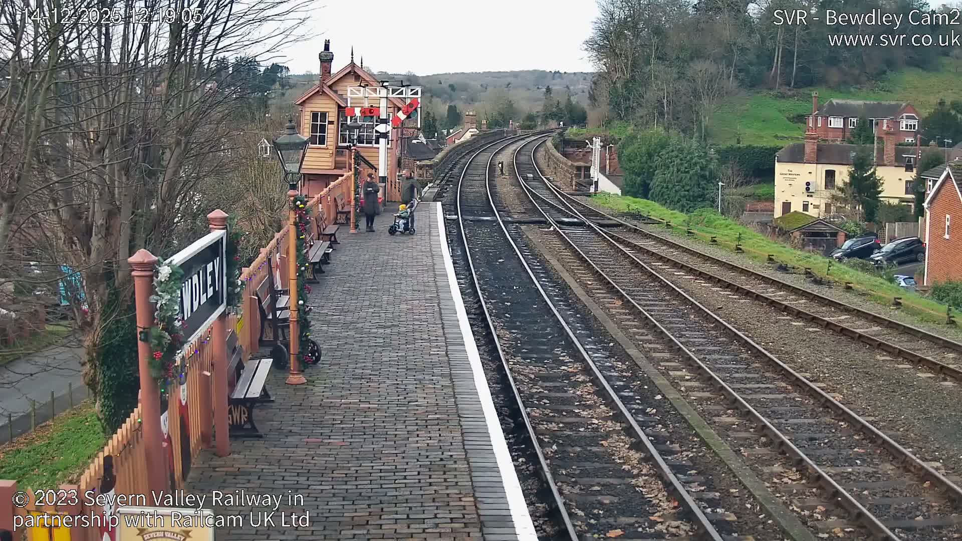 A charming old-fashioned railway station platform, adorned with festive garlands and benches, stretches alongside multiple frosty tracks that curve into a misty, tree-lined landscape under a cold, hazy sky.