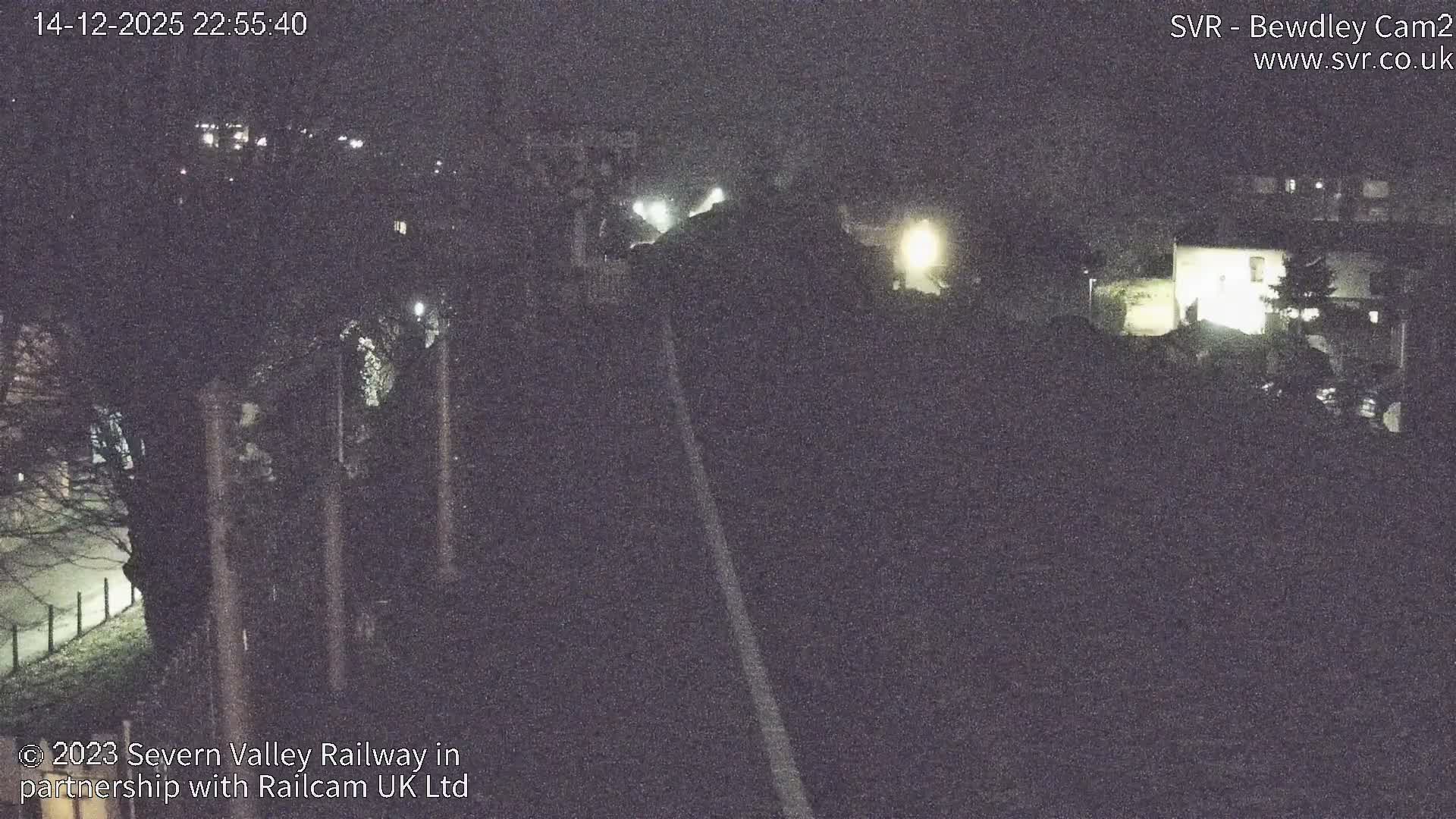 A charming old-fashioned railway station platform, adorned with festive garlands and benches, stretches alongside multiple frosty tracks that curve into a misty, tree-lined landscape under a cold, hazy sky.