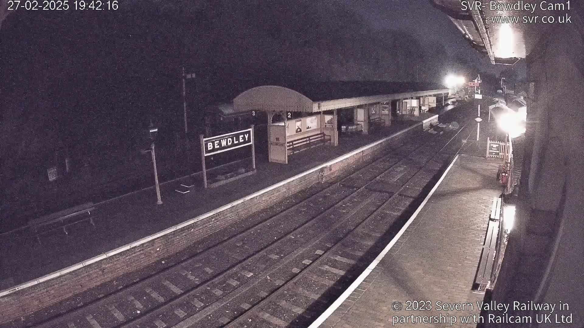 A nighttime view of a train station platform with train tracks and a partially visible train.
