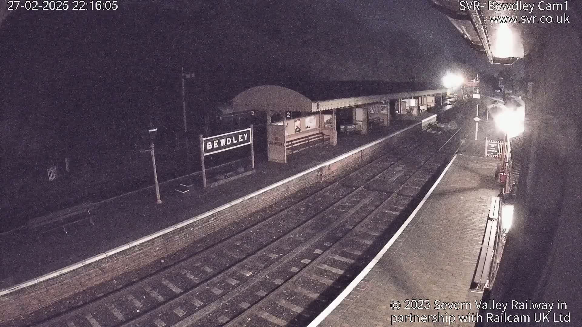 A nighttime view of a train station platform with train tracks and a sign reading "Bewdley."