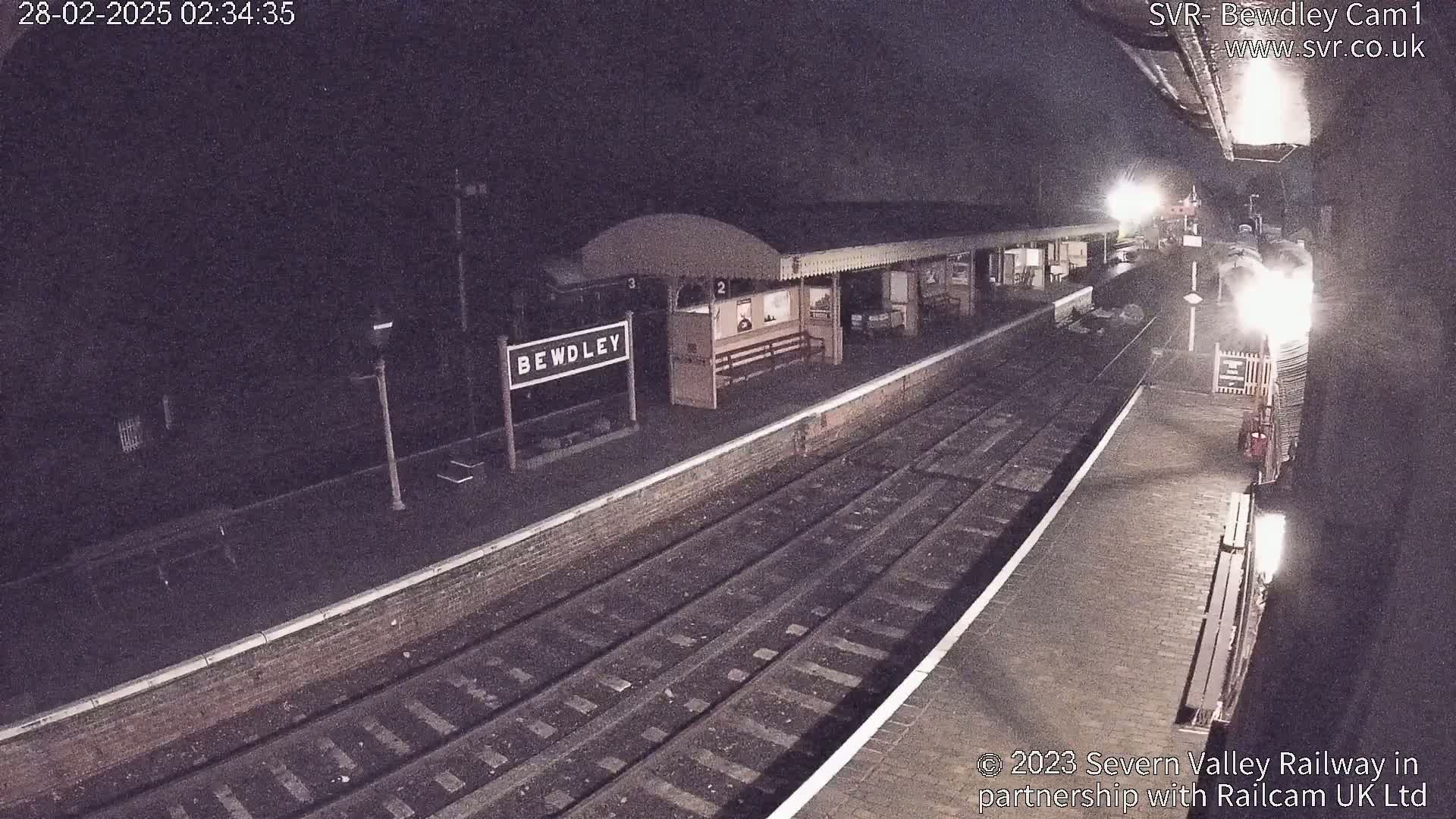 A nighttime view of a train station platform with a train arriving, under dark skies.