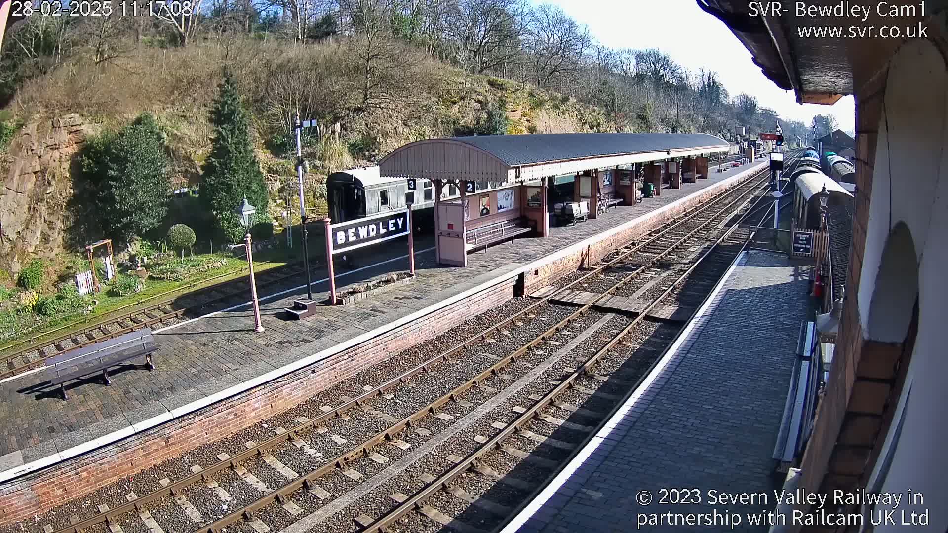 A train station platform with a covered waiting area, railway tracks, and a train partially visible on a sunny day.