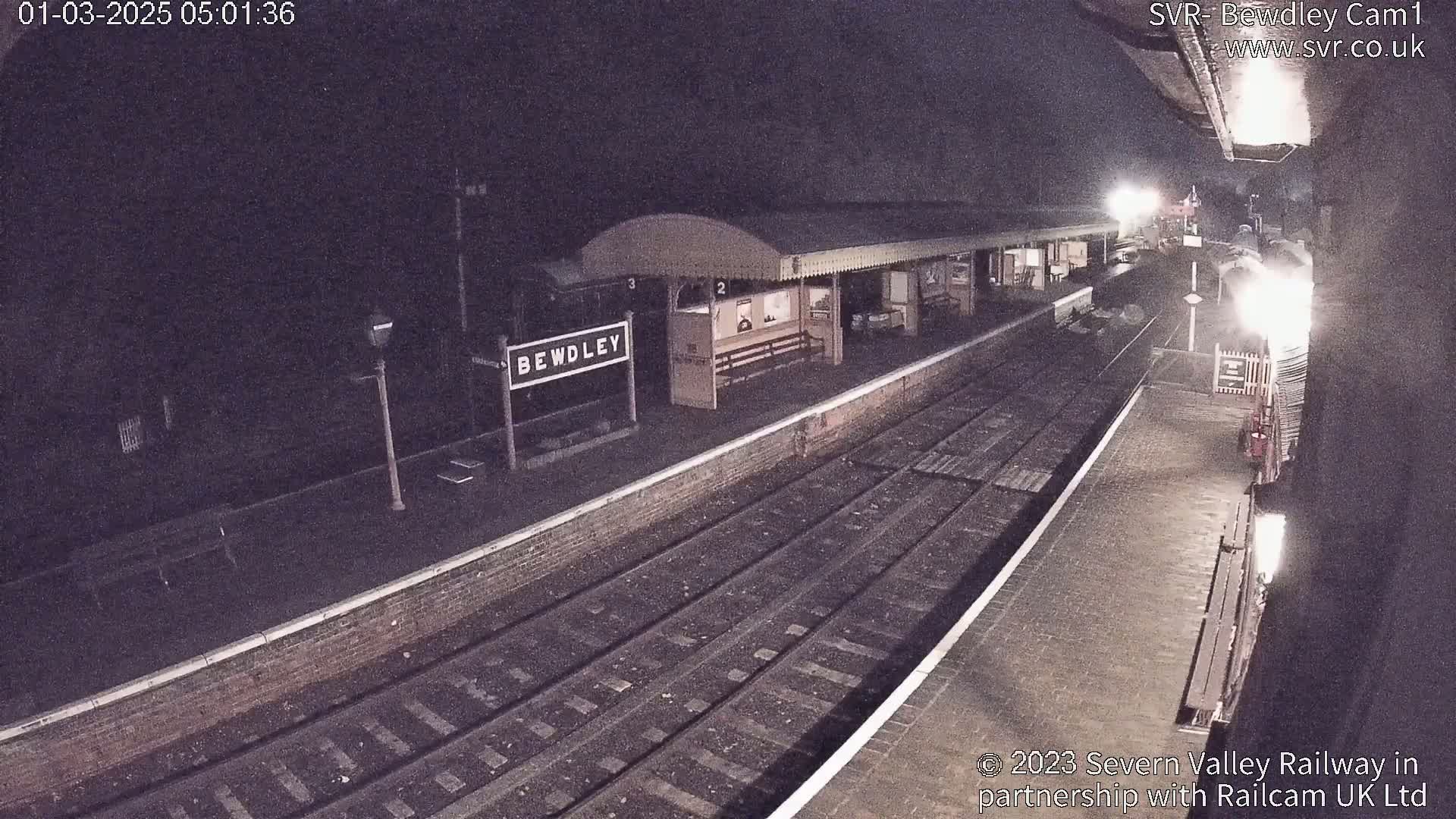 A nighttime view of a train station platform with train tracks and a partially visible train at the end of the platform.