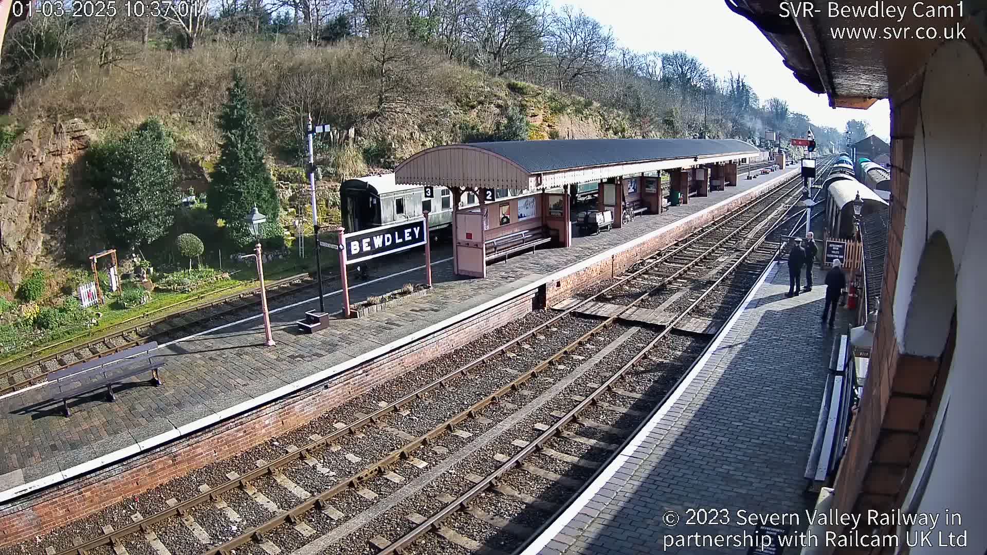 A sunny day at a train station with a covered platform, several train cars, and a few people standing on the platform.